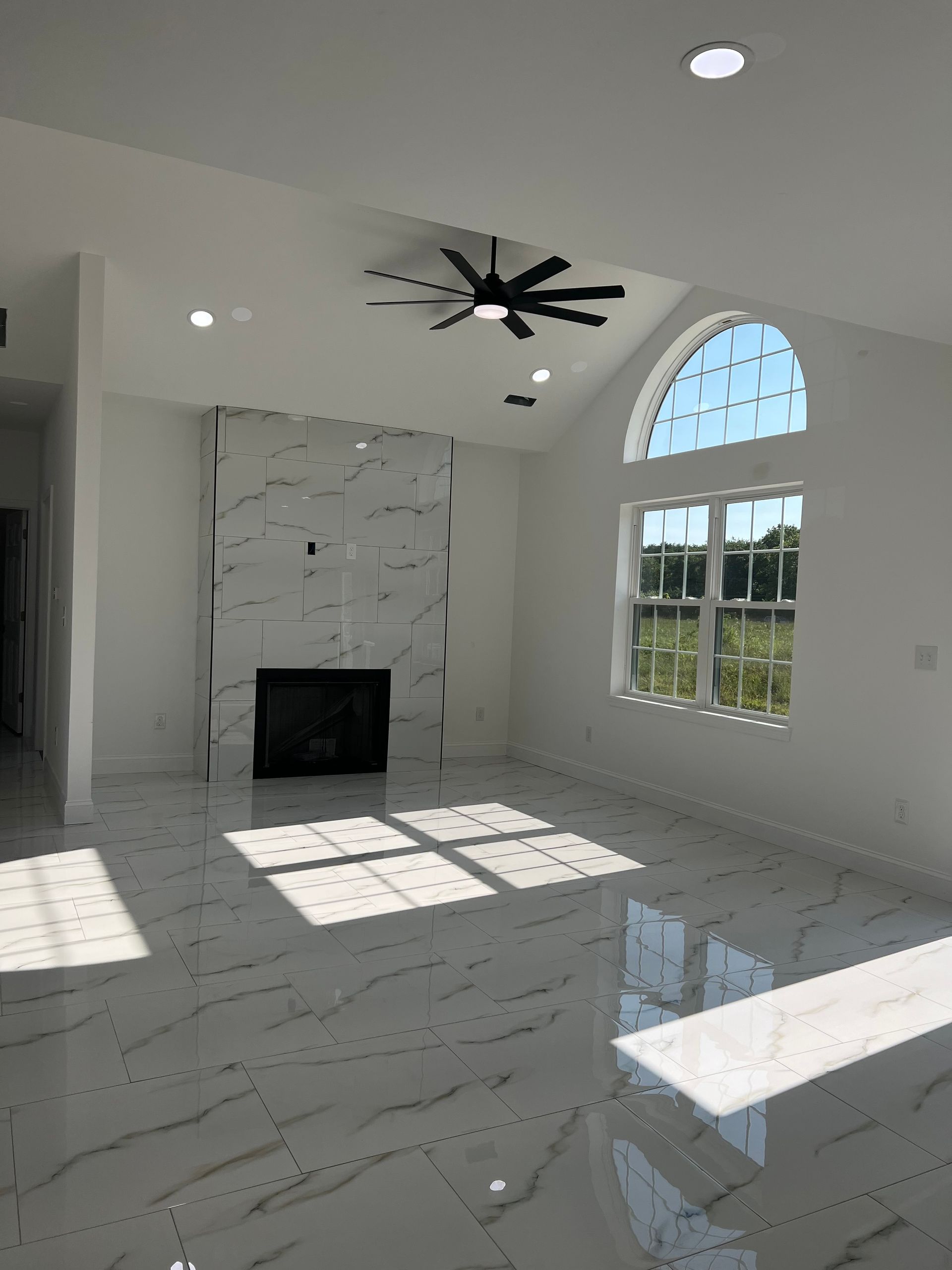 Bright modern living room with marble fireplace, arched window, ceiling fan, and sunlight on glossy floor