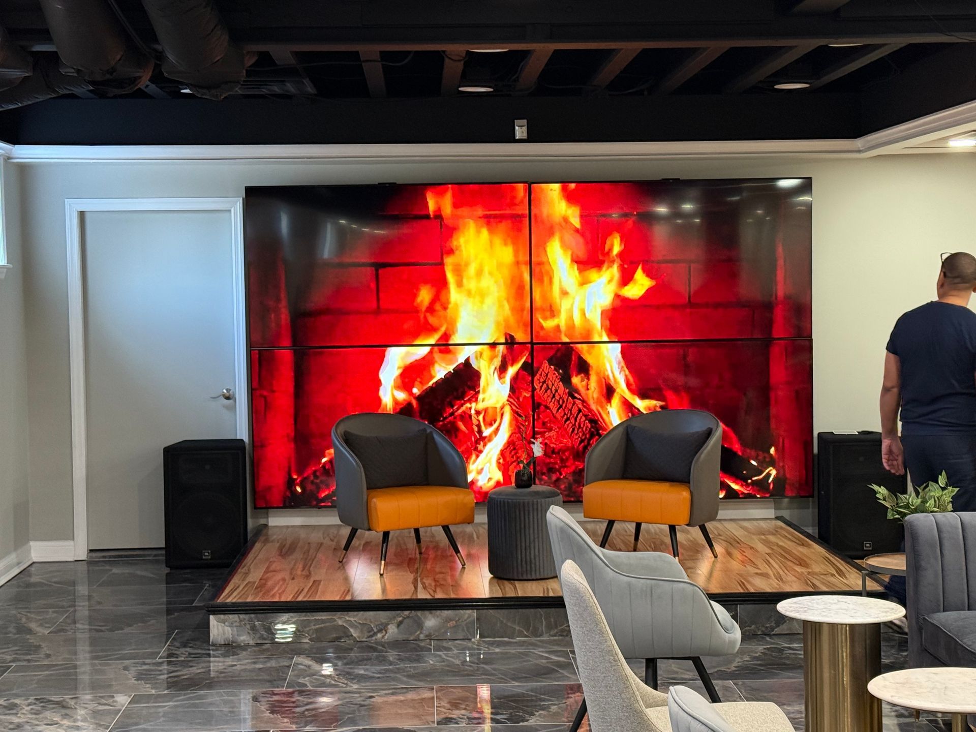 Lounge with wall-sized fireplace screen showing a roaring fire, chairs, rug, and a person standing at right