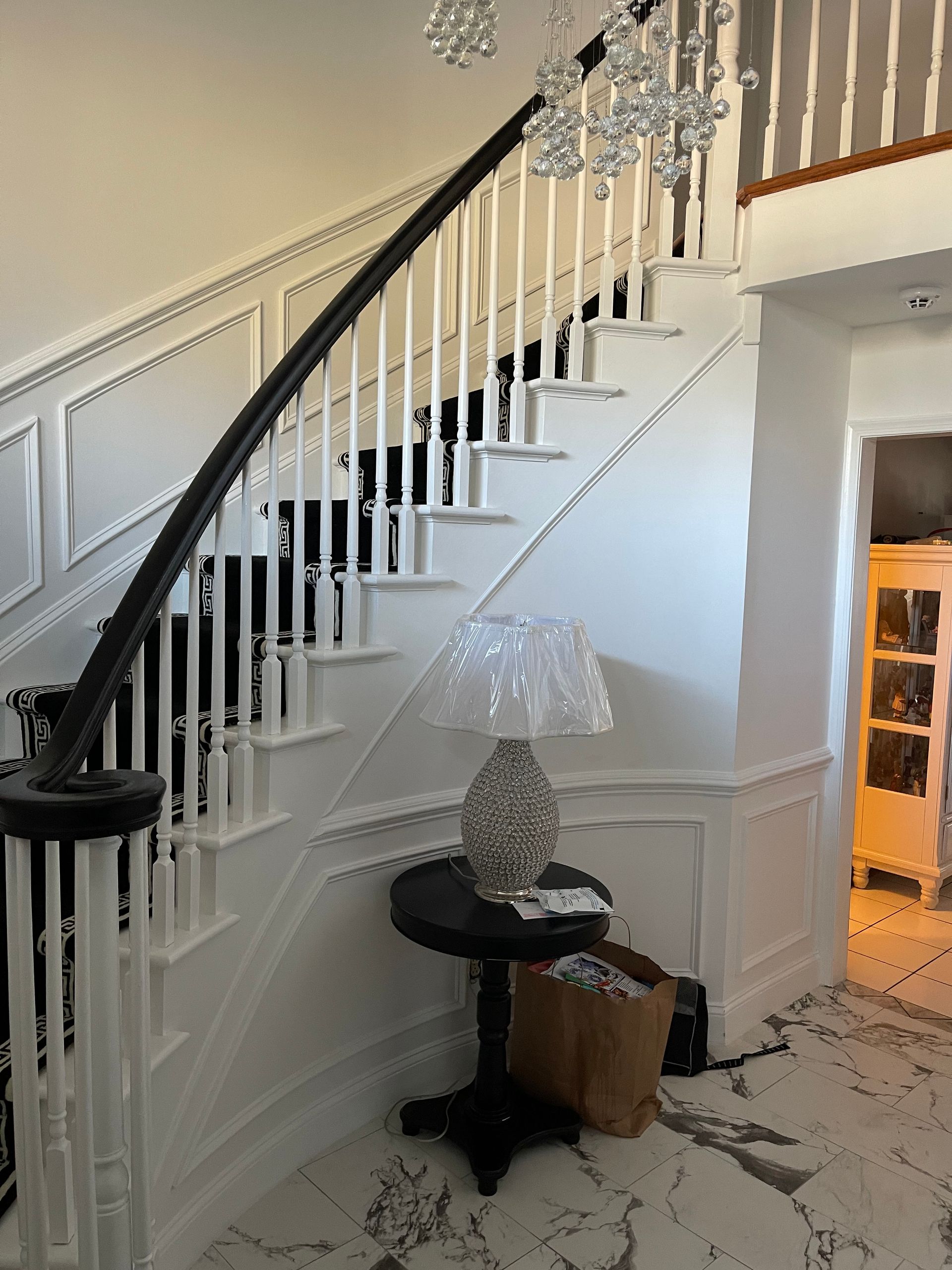 Curved staircase with white railings, black handrail, crystal chandelier, and small table lamp in a foyer