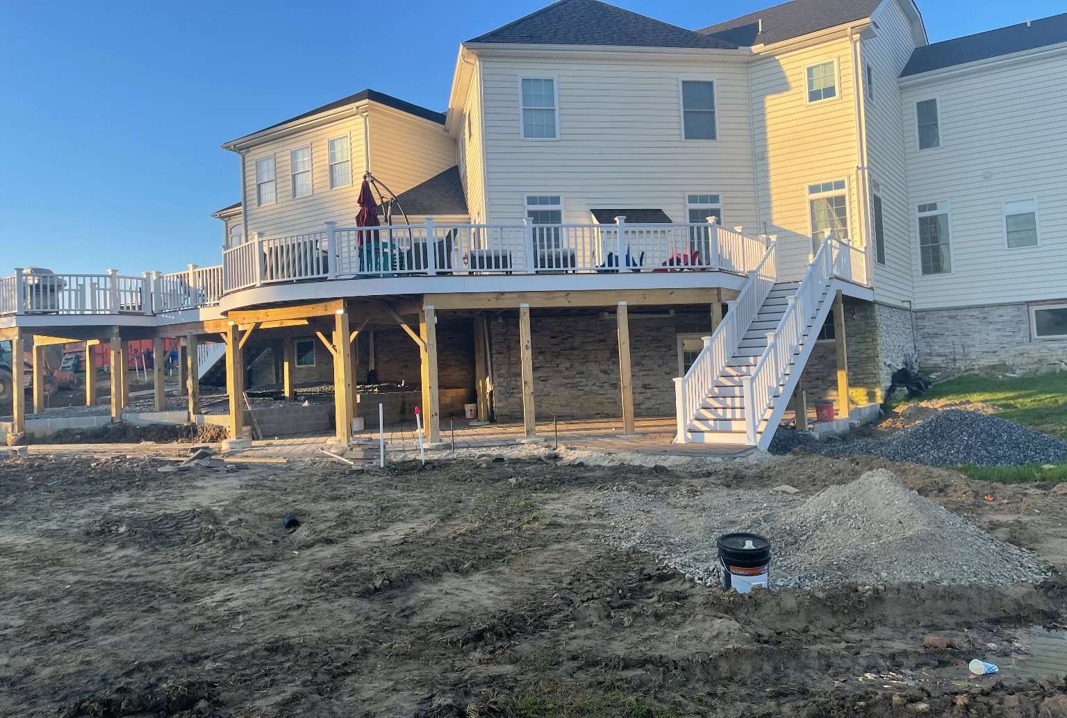A large light-colored house with a recently built wooden deck, white railing, and stairs leading to an unfinished yard