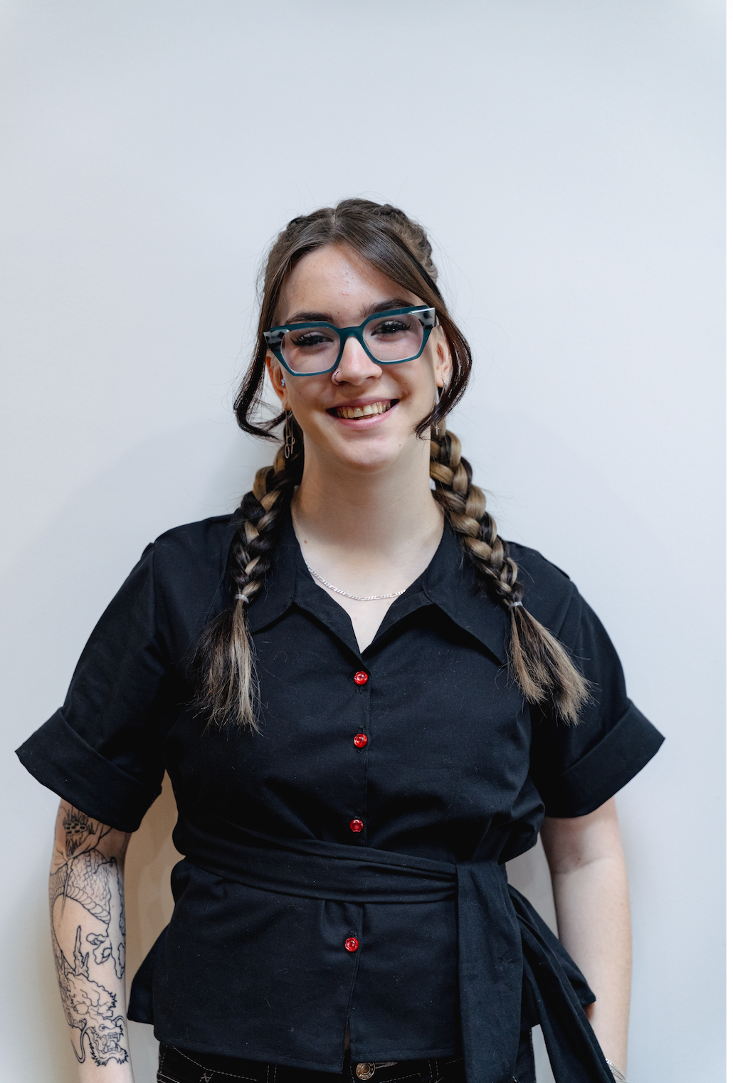 Person standing against a light background, smiling in a black shirt with braided hair and glasses.