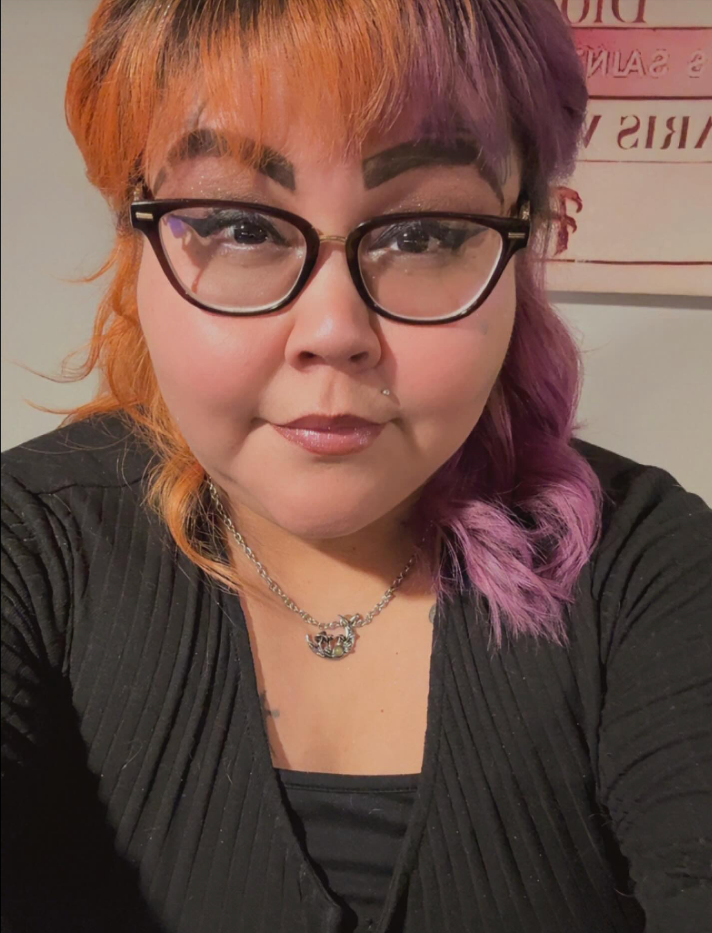 Close-up selfie of a person with orange and purple hair, glasses, and a black top, looking at the camera.