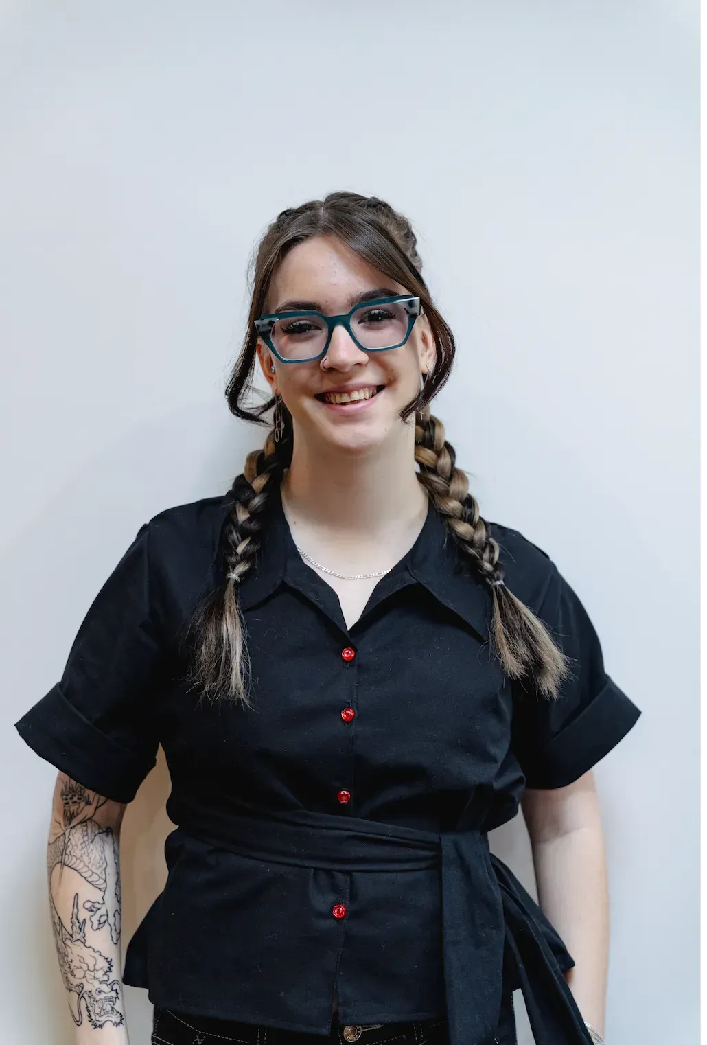 Person standing against a light background, smiling in a black shirt with braided hair and glasses.