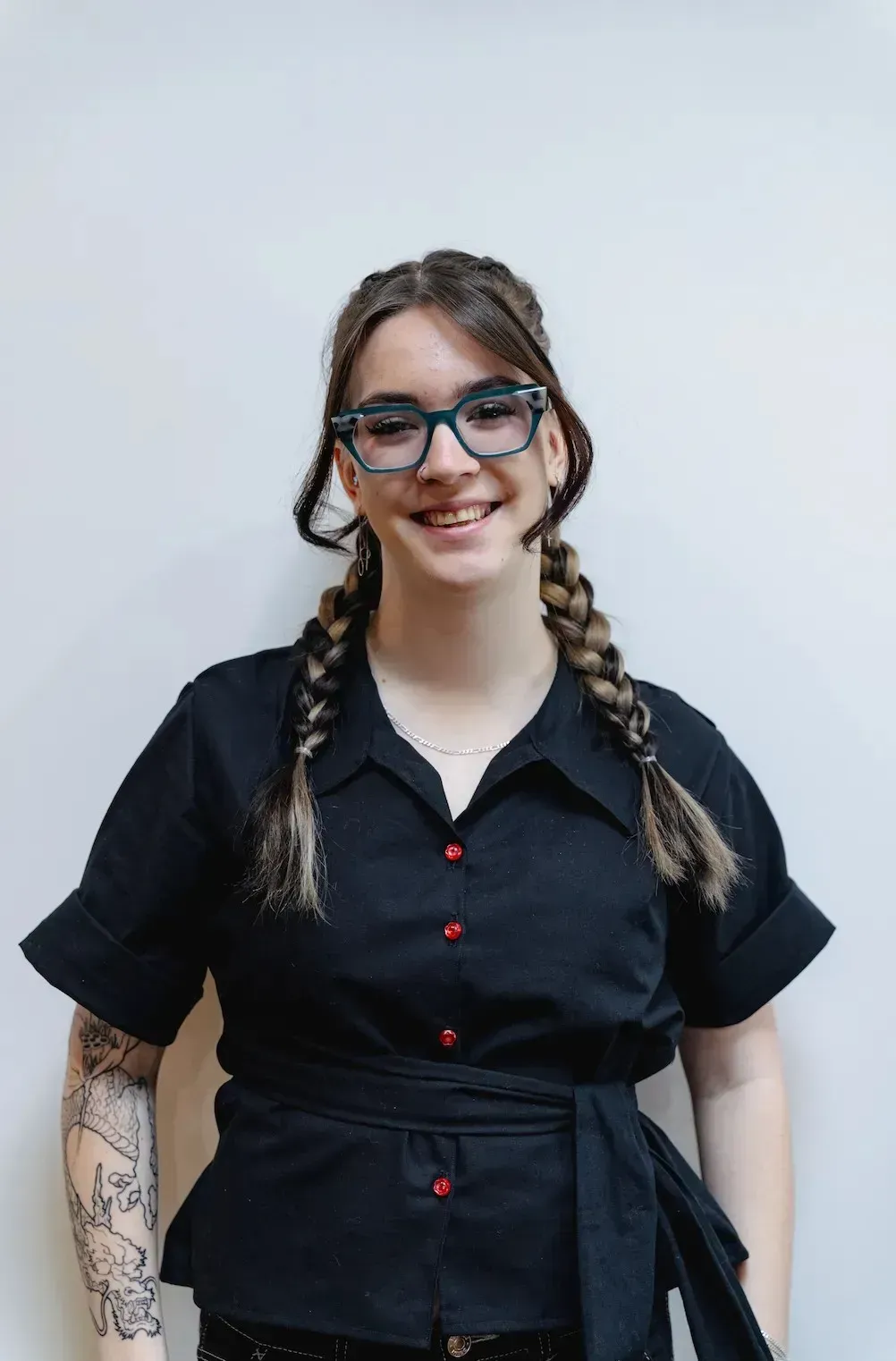 Person standing against a light background, smiling in a black shirt with braided hair and glasses.
