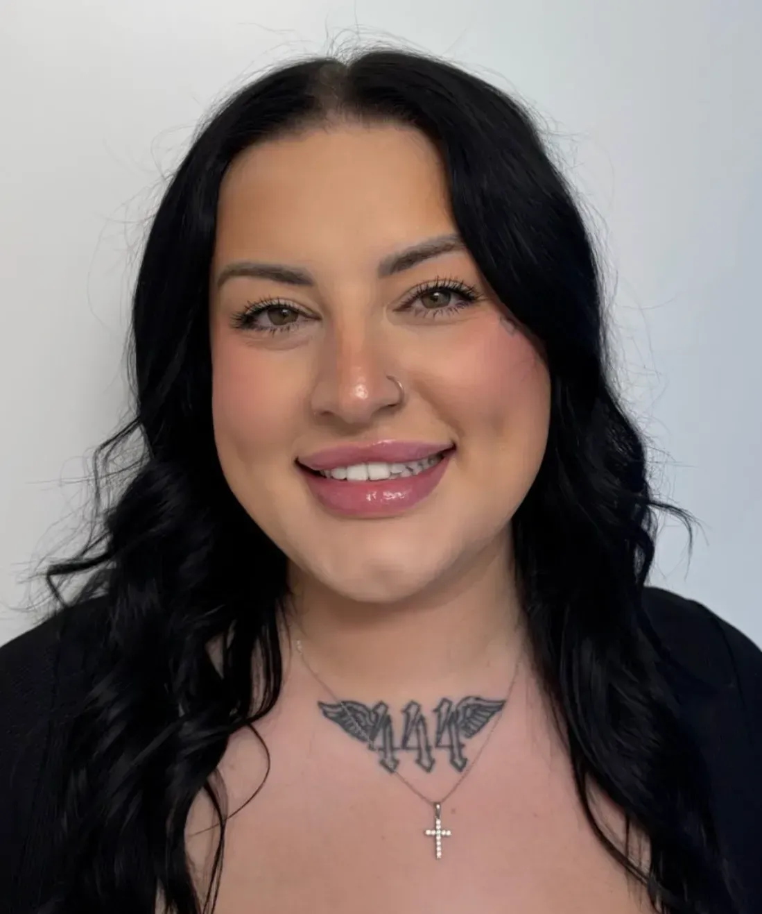 Smiling woman with dark hair, nose ring, chest tattoos, and black top against a light background