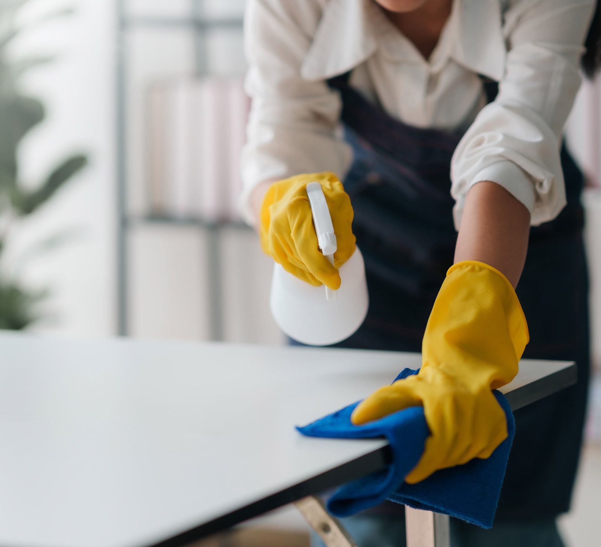 A woman wearing yellow gloves is cleaning a table with a cloth and spray bottle.