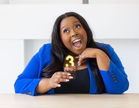 Anisha Angella, Founder and CEO, The Early Childhood Coach smiling at her desk holding a cupcake as she celebrates her third year as an ECE coach