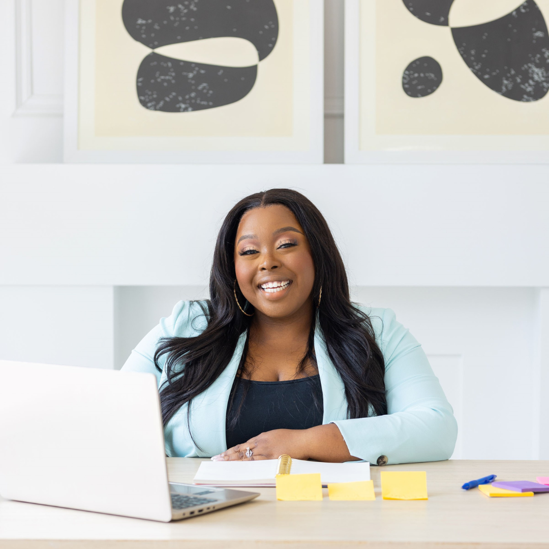 Anisha Angella, the Early Childhood Coach, posing and smiling in front of her desk with a hand on her hip and holding her laptop