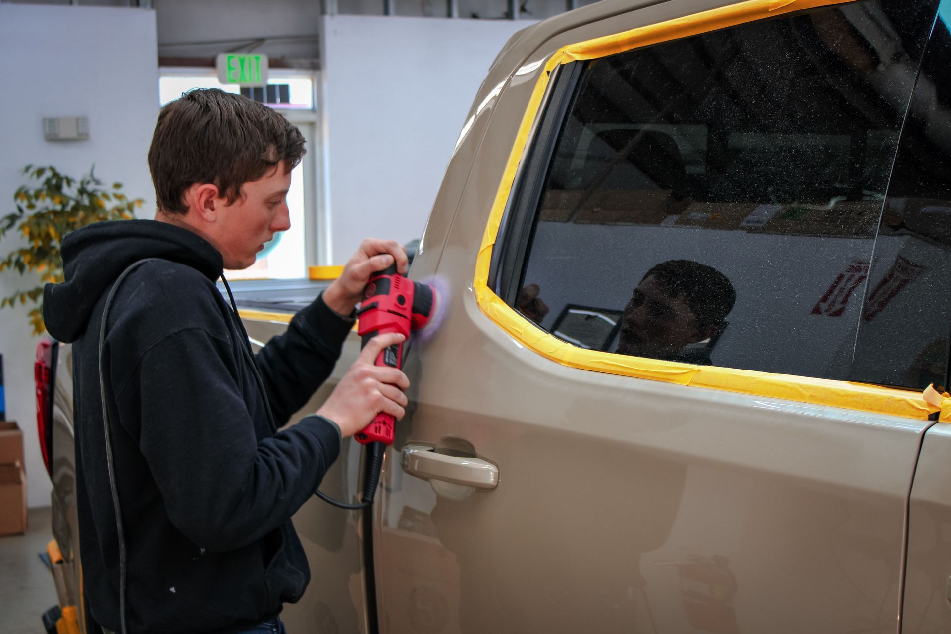 Man using a polisher on a tan car in a garage. Yellow tape protects the windows.