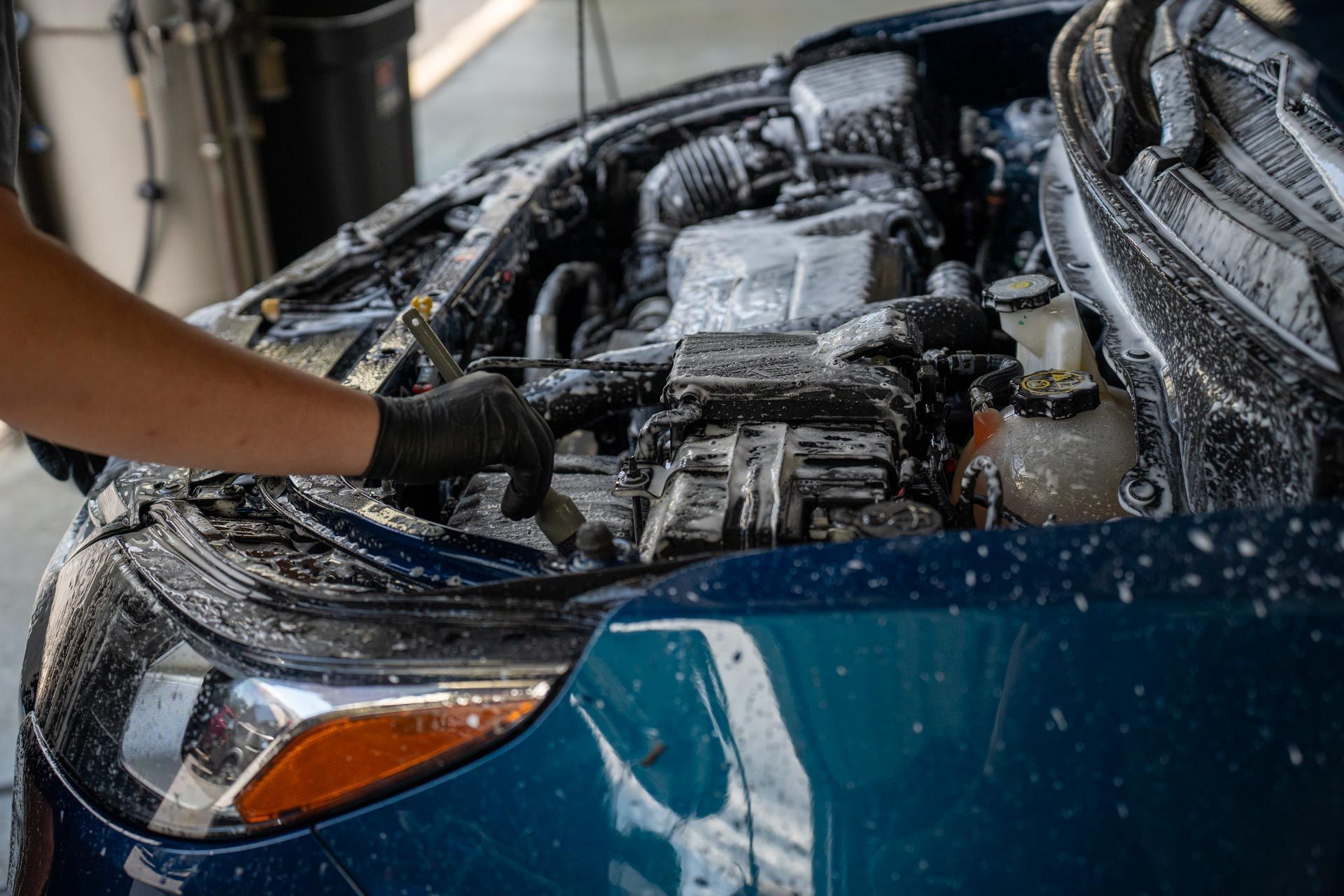 A person cleaning a car engine with foam and a brush. Blue car in a garage.