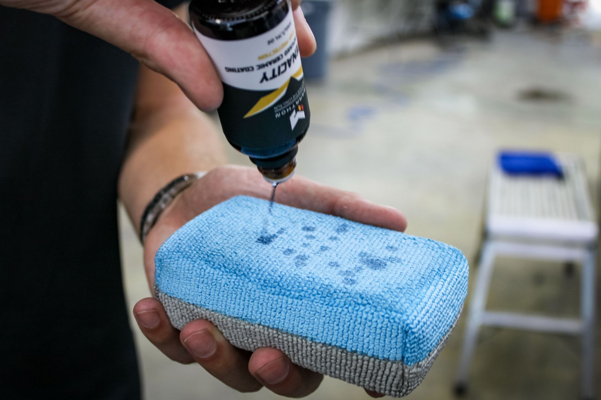 Person dispensing liquid from a bottle onto a blue sponge, likely for detailing a vehicle.