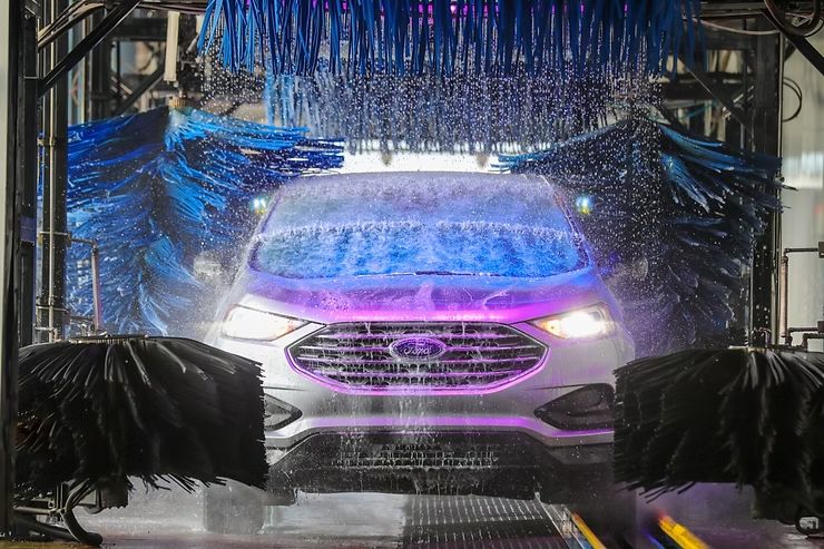 White Ford SUV in a car wash, being sprayed with water and scrubbed by rotating blue and black brushes.