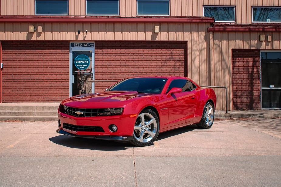 A bright red Chevrolet Camaro parked in front of a tan brick building with metal siding and glass entry doors.