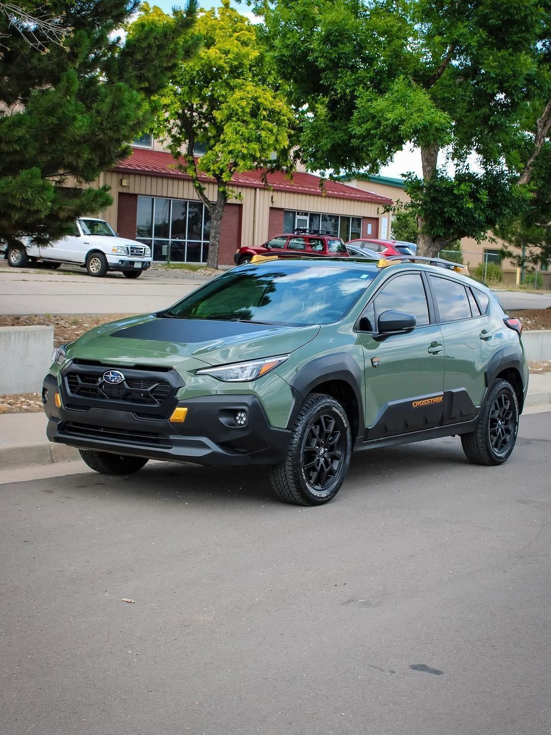 Green Subaru Crosstrek parked on asphalt. Black accents, black wheels, against a backdrop of trees and a building.