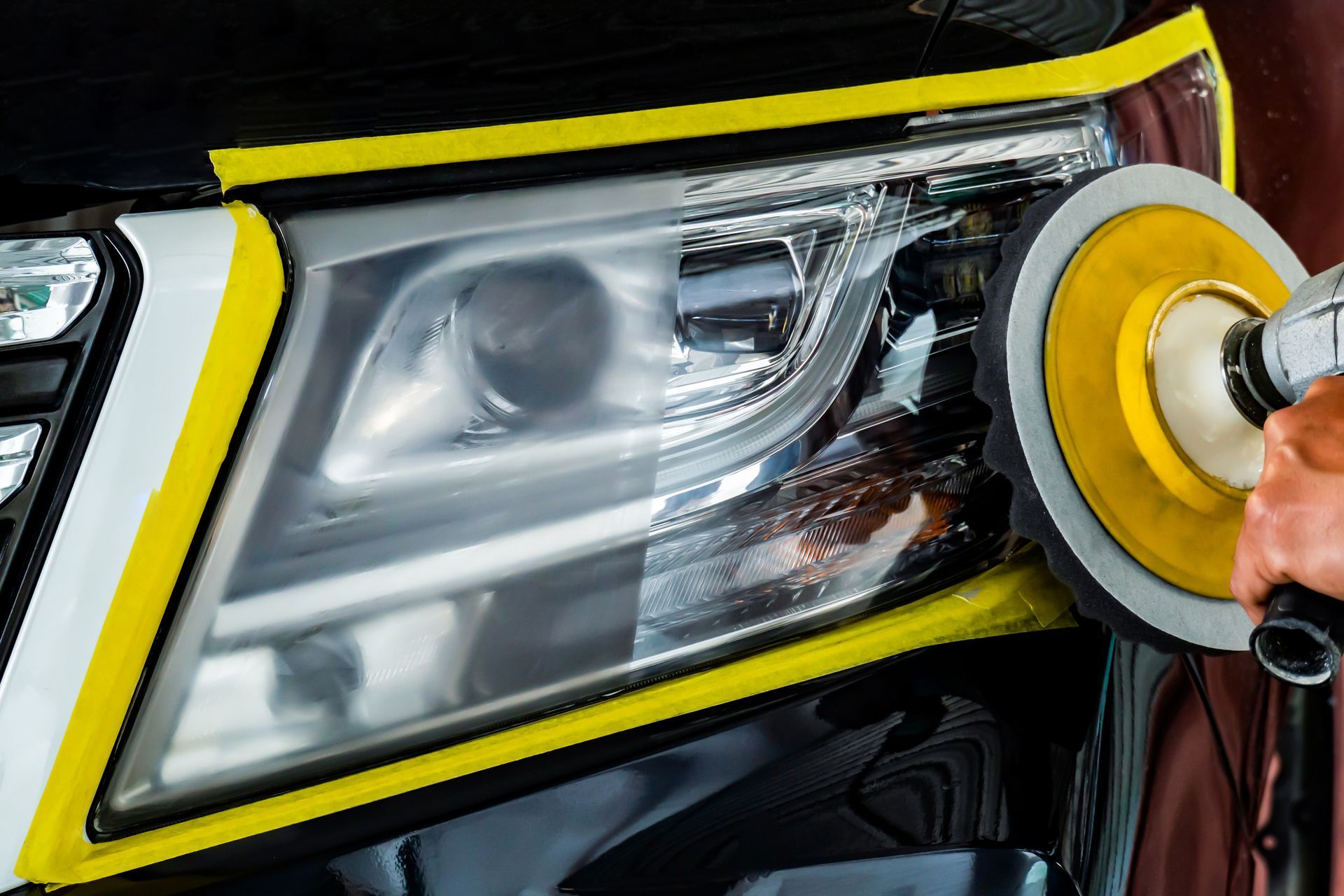 Headlight polishing: a person uses a power buffer on a car's headlight. The left side is clear, the right is hazy.