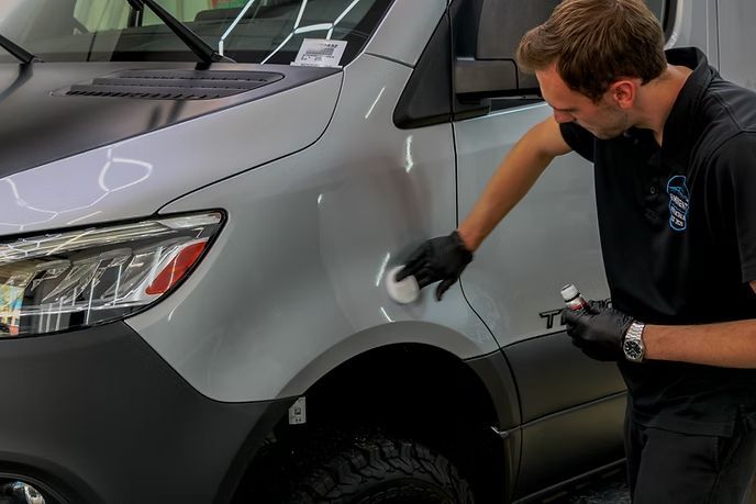 A person wearing black gloves applies a ceramic coating to the side panel of a gray van in a garage.