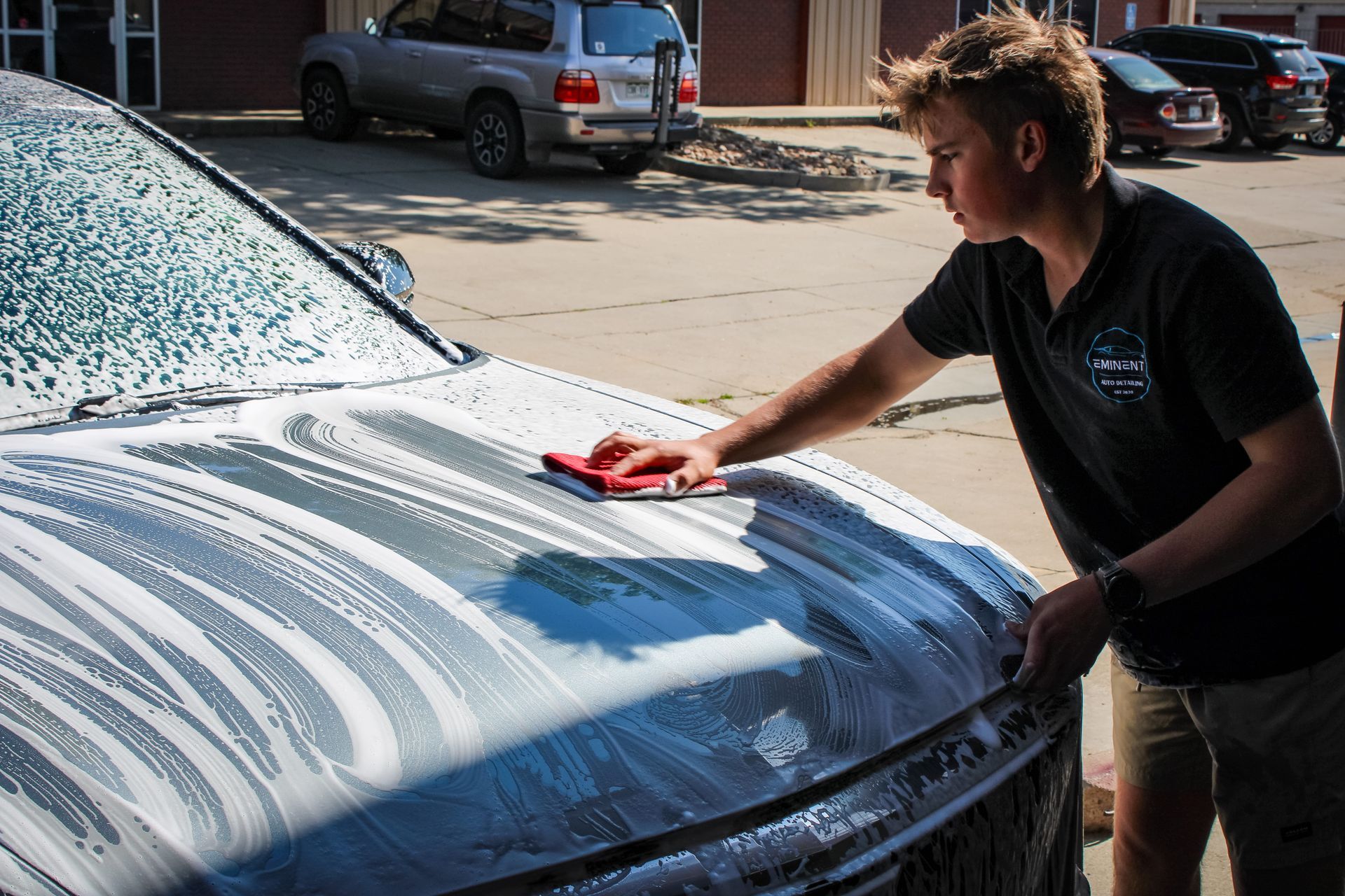 Person washes a car hood with soap and a red sponge outside.