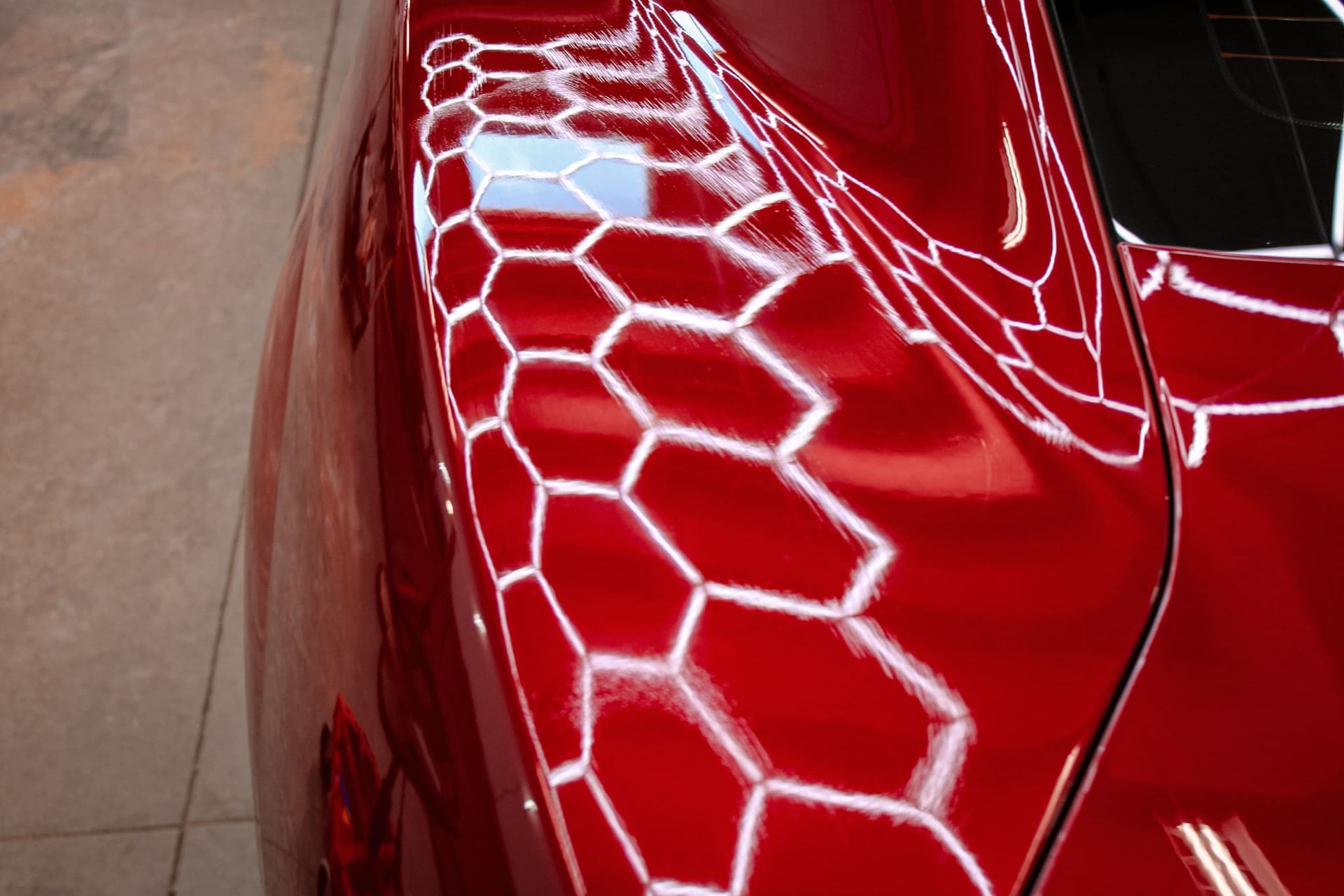 Red car's glossy surface reflecting a grid of hexagonal shapes, likely from overhead lights.