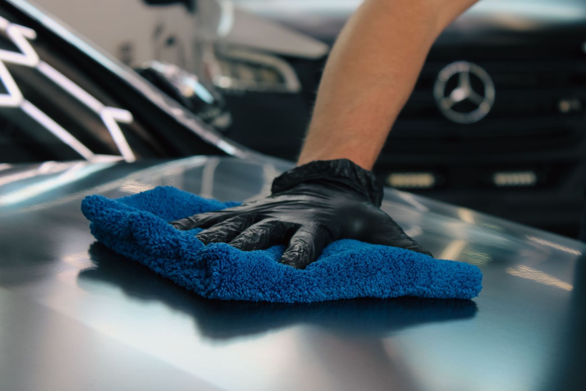Hand in black glove wipes a blue microfiber towel across the hood of a car