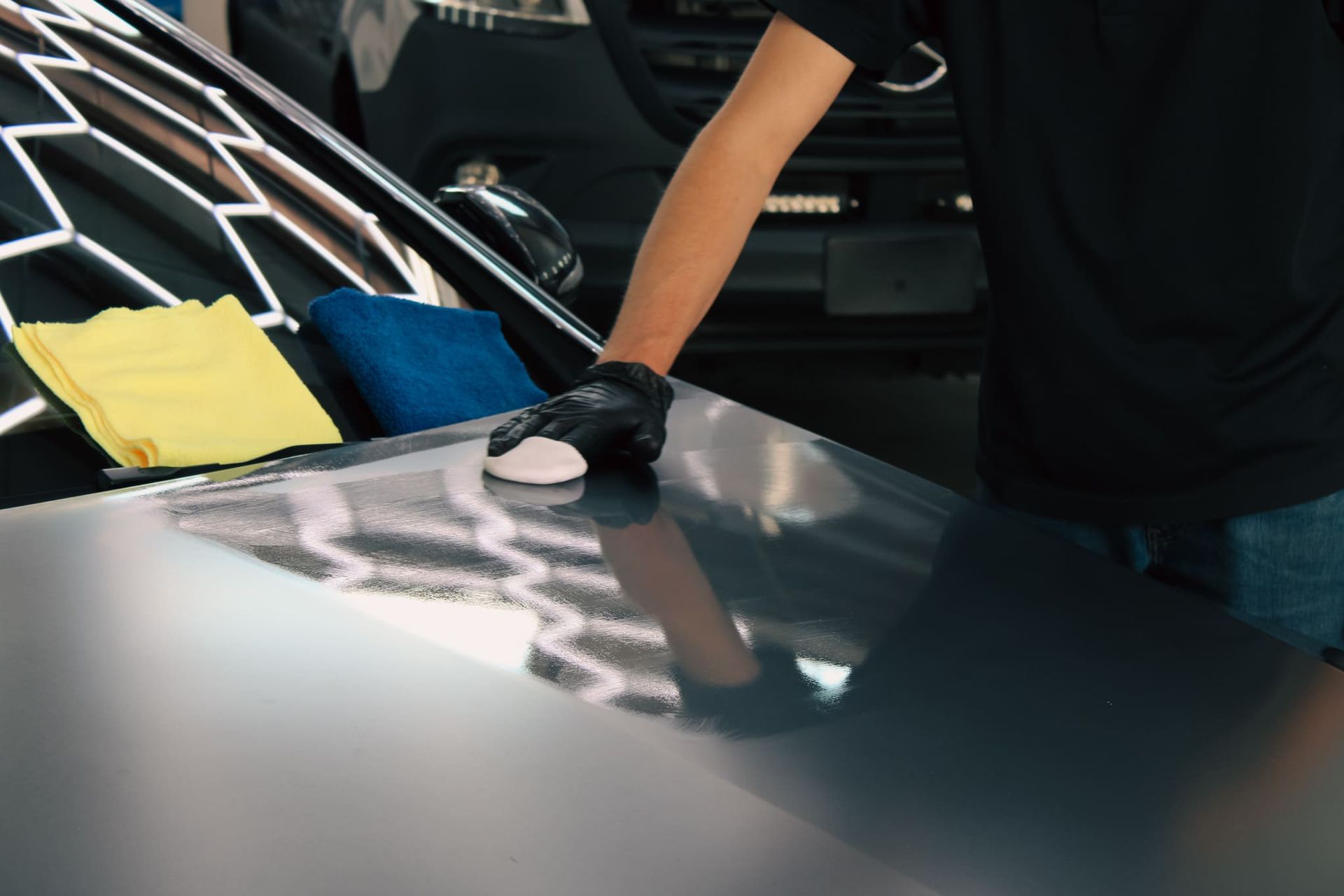 Person wearing gloves polishing a car hood; yellow and blue cloths are nearby.