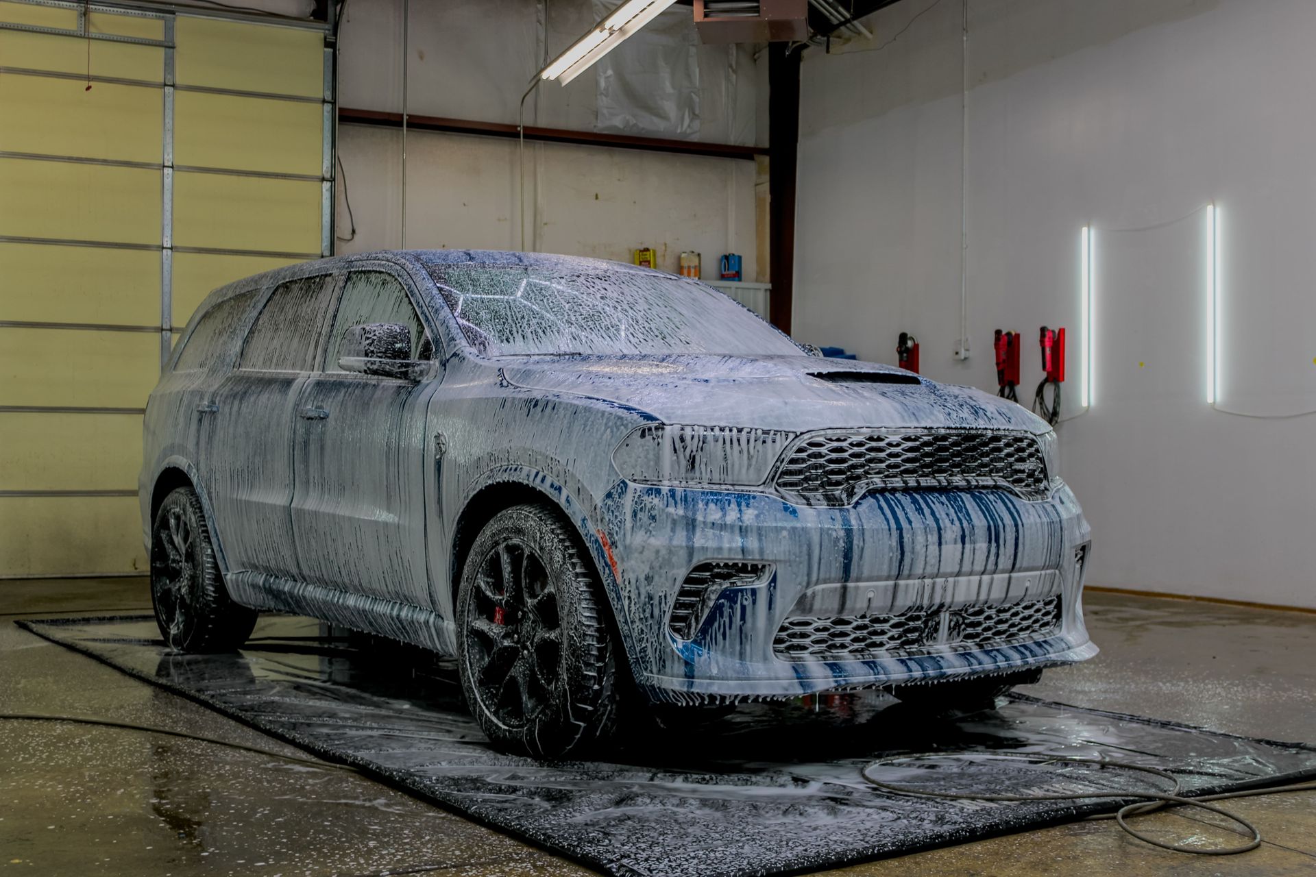 Blue SUV covered in white foam at a car wash, sitting on a black mat indoors.