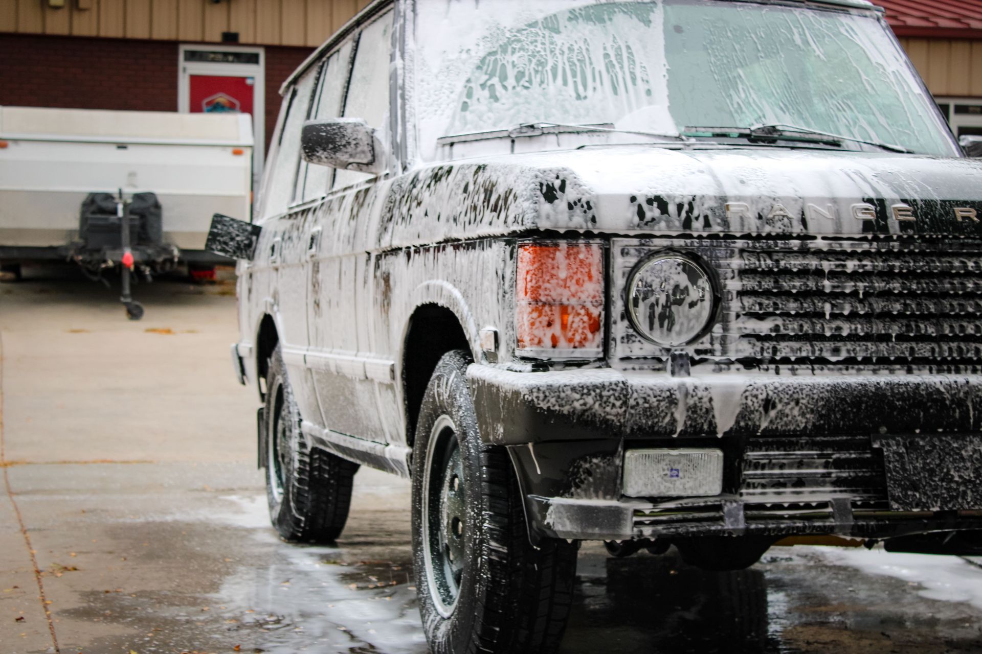 Black SUV covered in soap suds, being washed in a driveway with a trailer in the background.