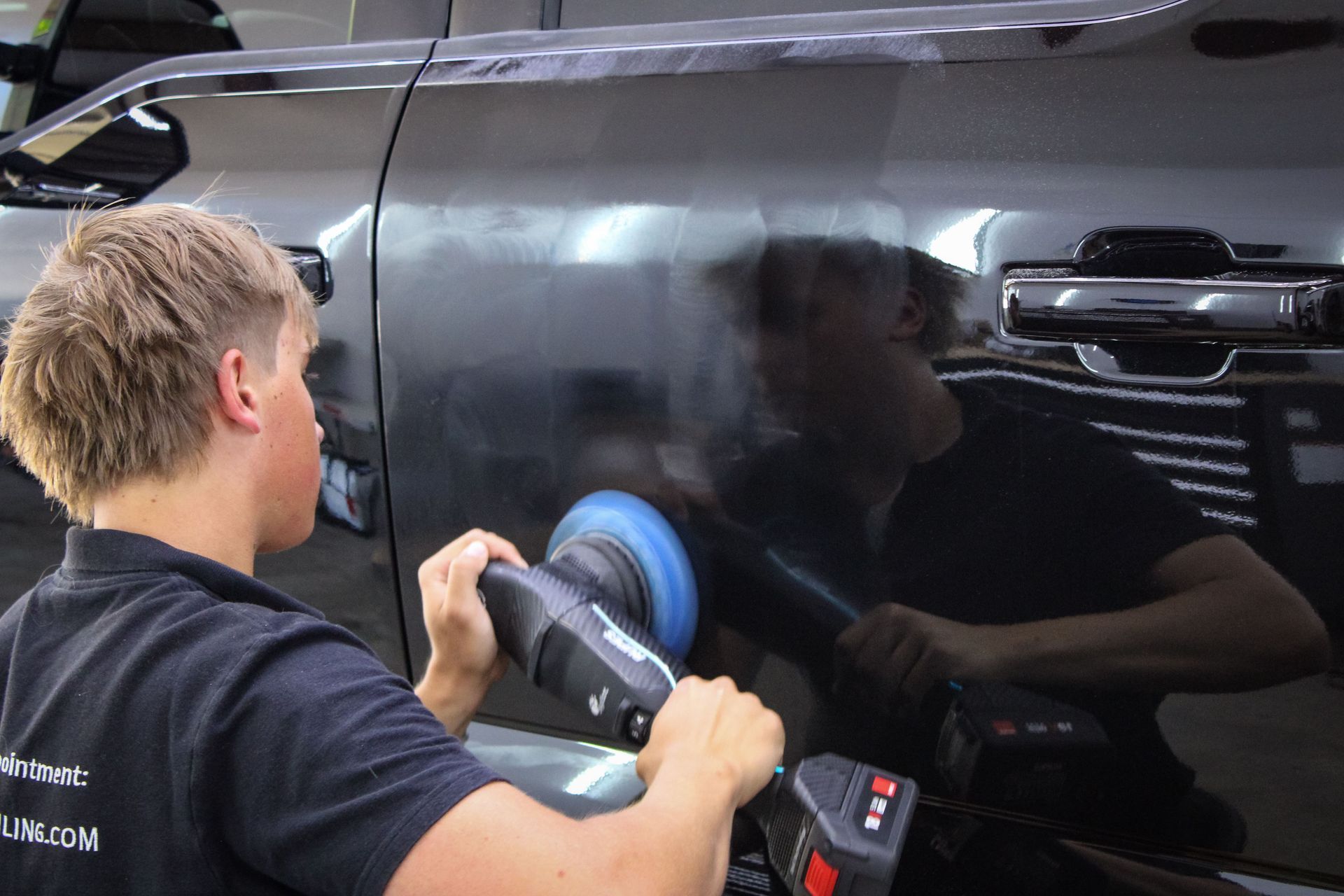Person polishing a black car with a power buffer in a well-lit shop.