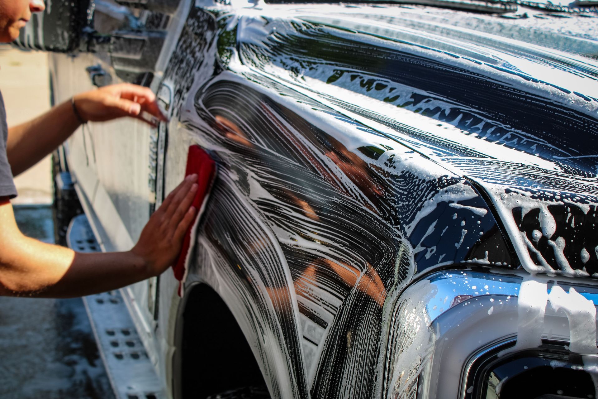 Person washing a black car with a red sponge, covered in white soap suds.