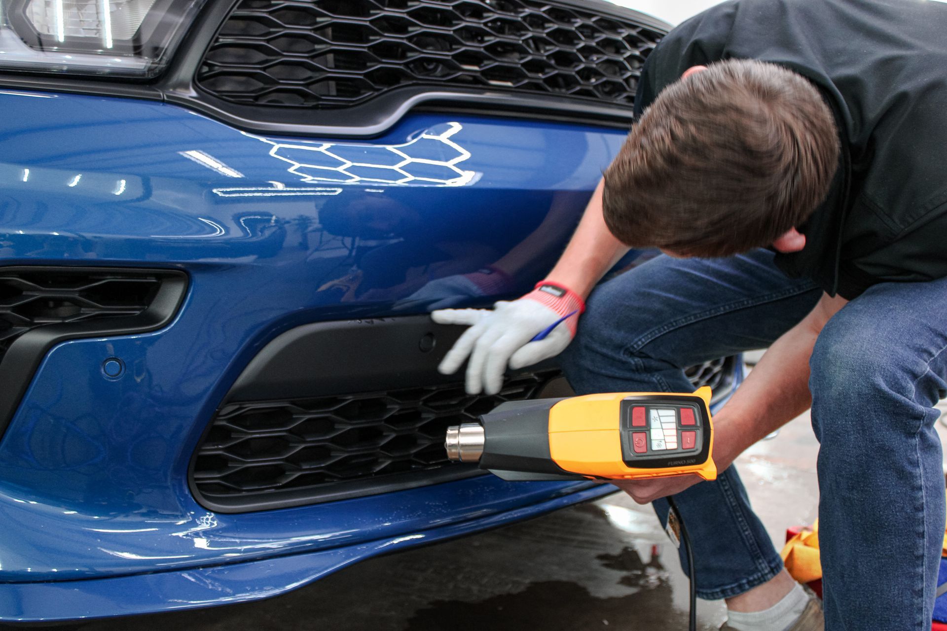 Man uses a heat gun on a car bumper in a shop. The car is blue.