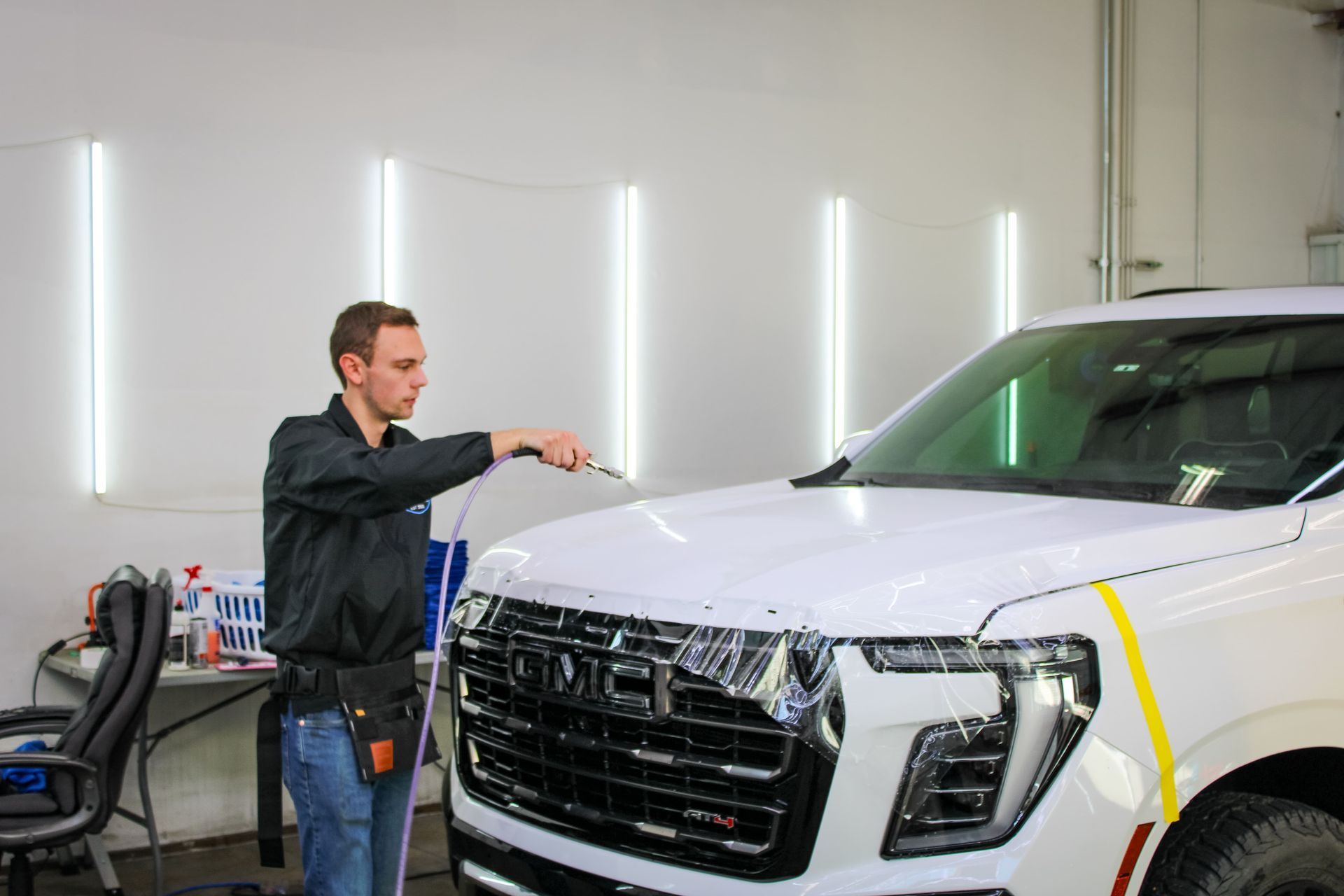 Man applying film to white truck hood in a shop. Fluorescent lights and tools visible.