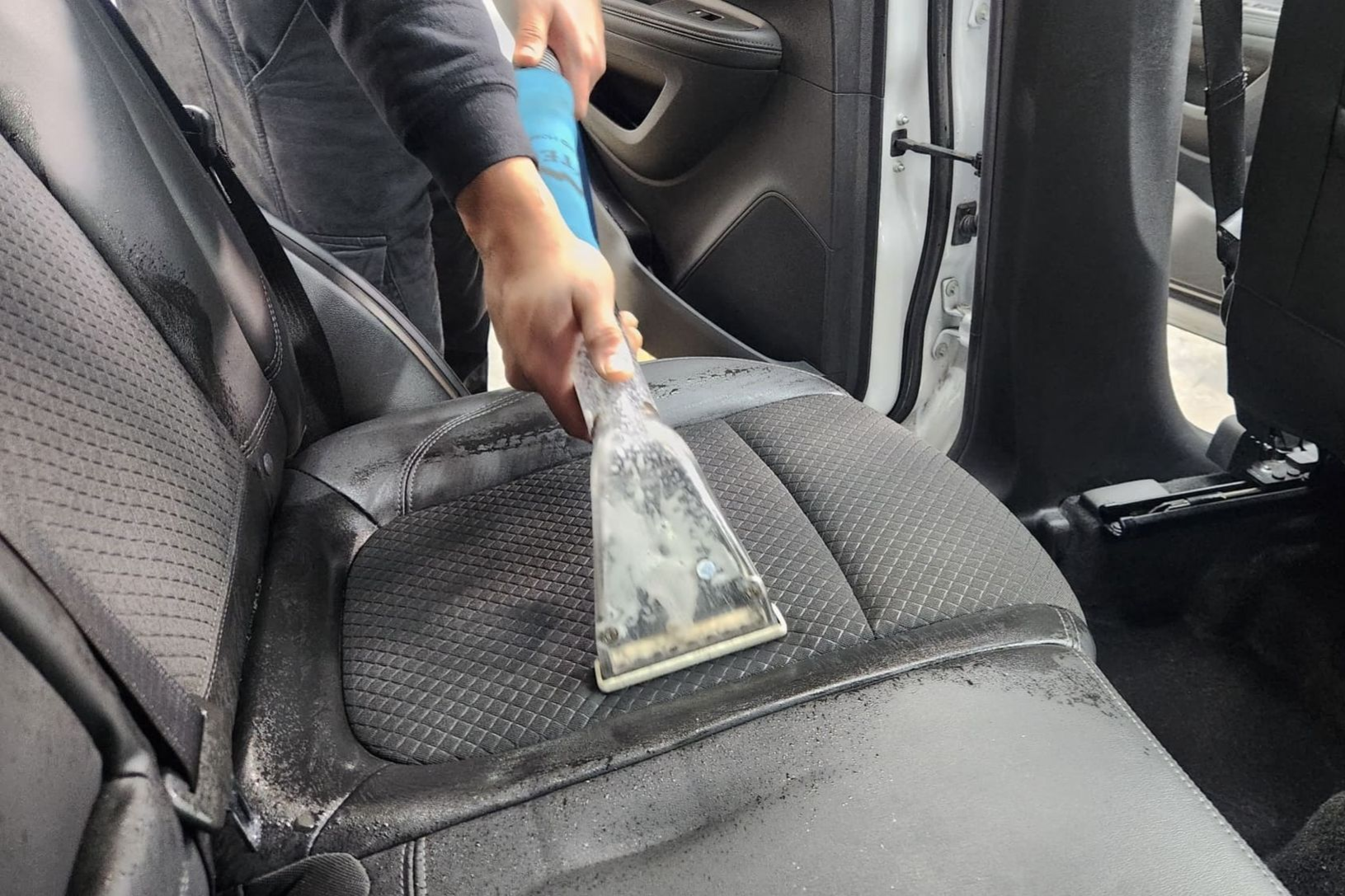 Person cleaning a black car seat with a handheld cleaning tool inside a white car.