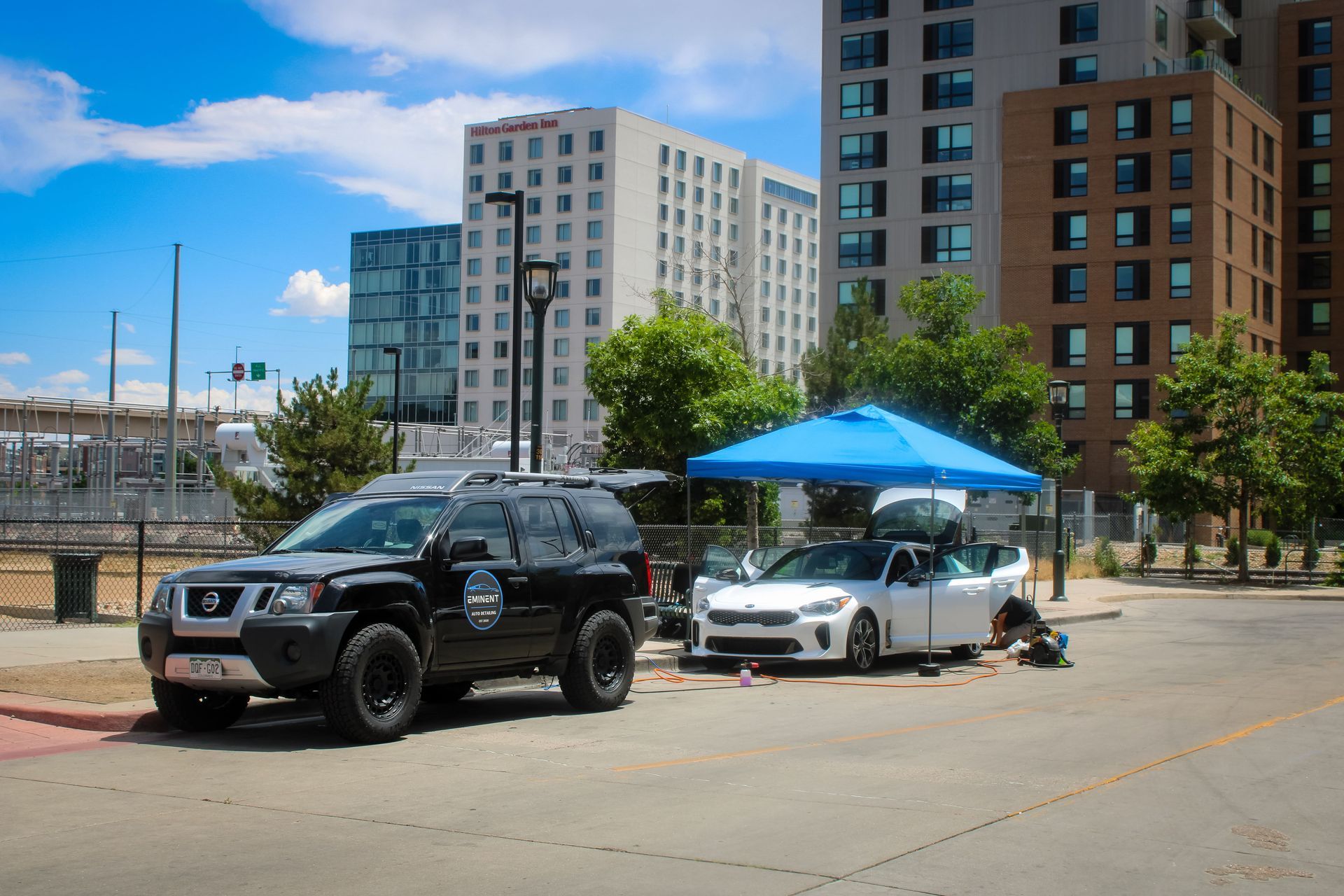 Two cars parked on a city street, one black SUV and one white car under a blue canopy. Buildings in background.