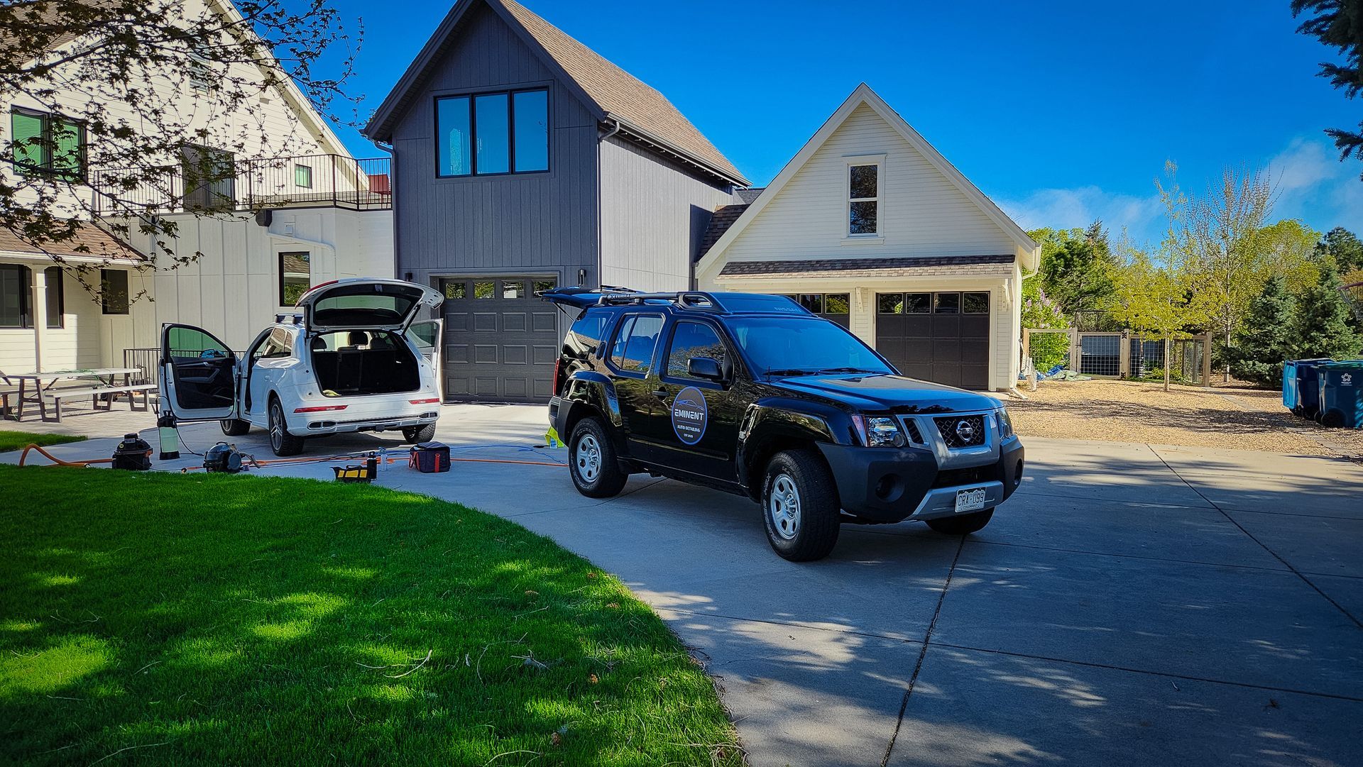Black SUV parked in driveway with garage and white car. Tools on ground. Houses in background.