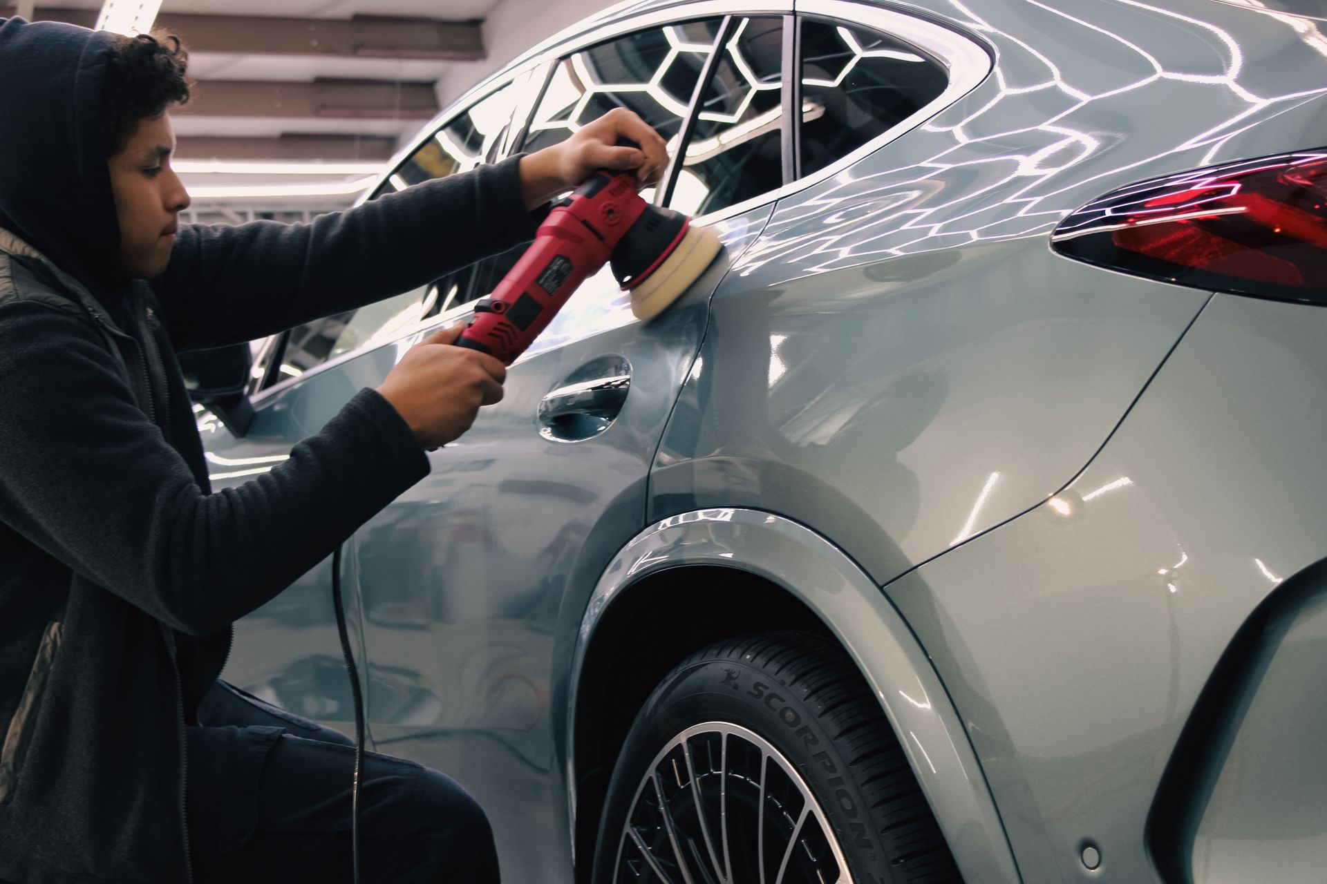 Person polishes a light-colored car with a power buffer. The person is in a well-lit workshop.
