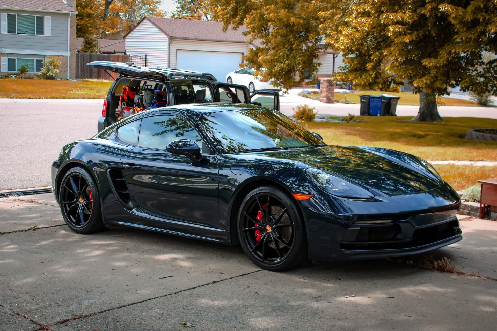 Dark blue Porsche sports car parked in a driveway. Tailgate open on a car behind. Red brake calipers visible.