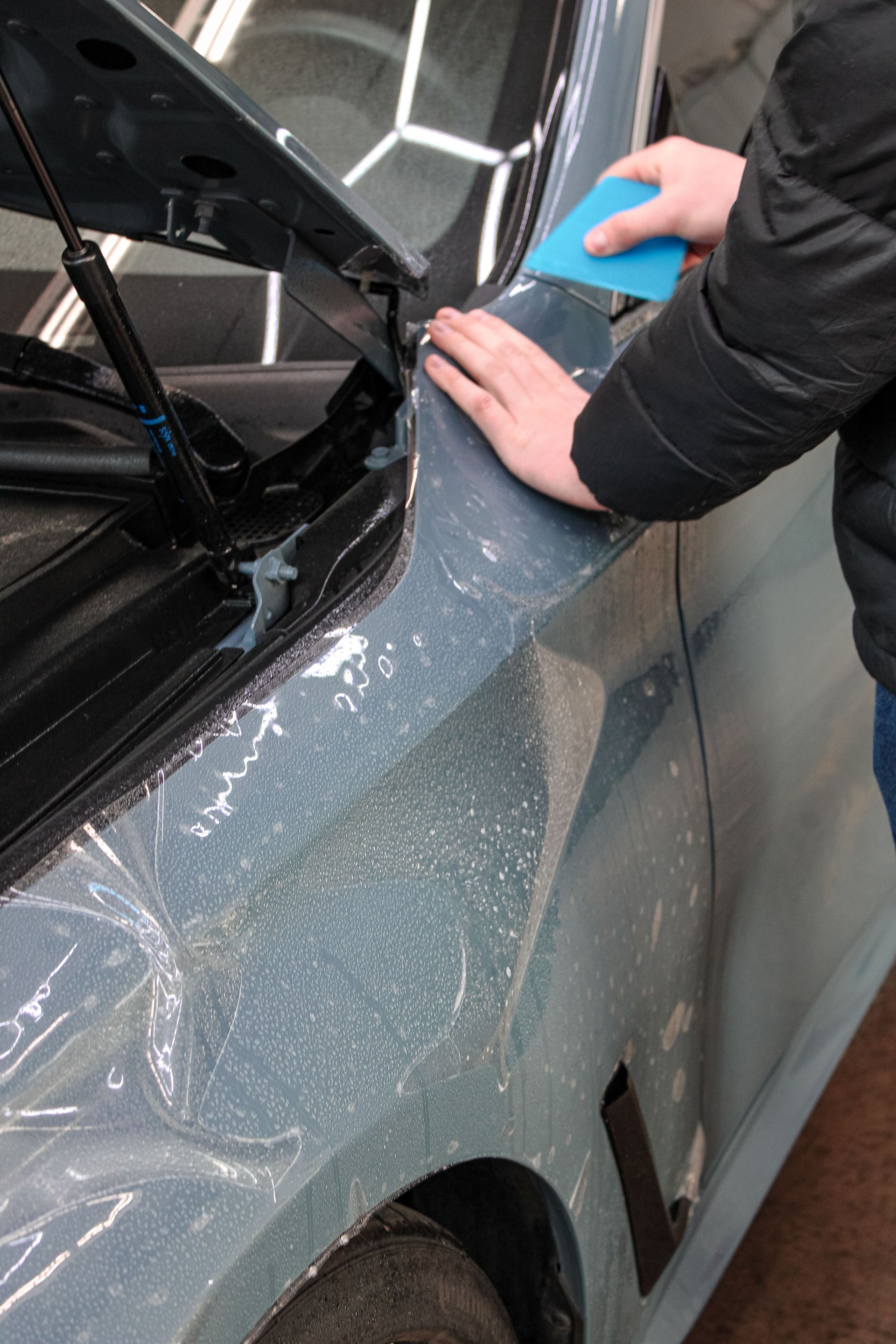 Man applying protective film to a black Cadillac SUV in a garage with detailing supplies.