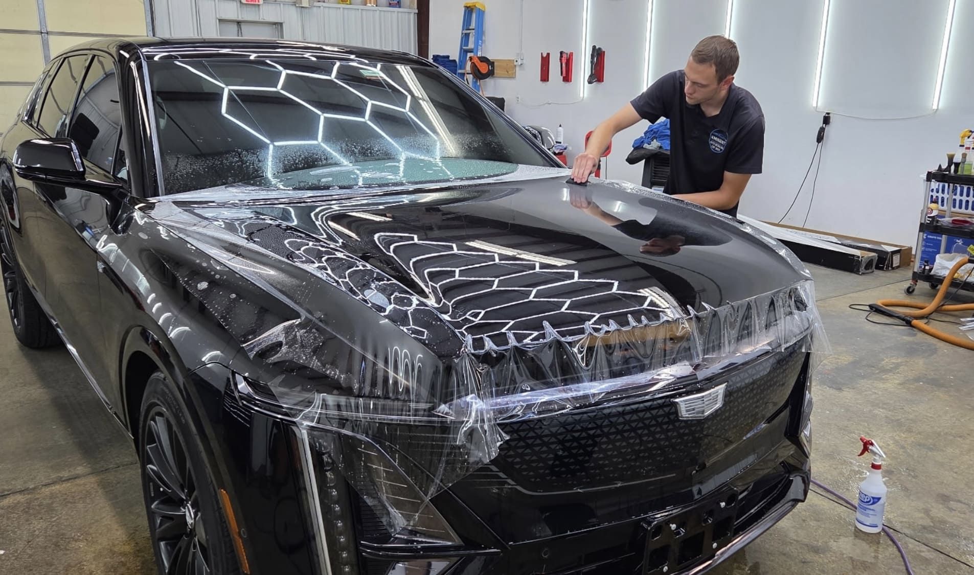Man applying protective film to a black Cadillac SUV in a garage with detailing supplies.