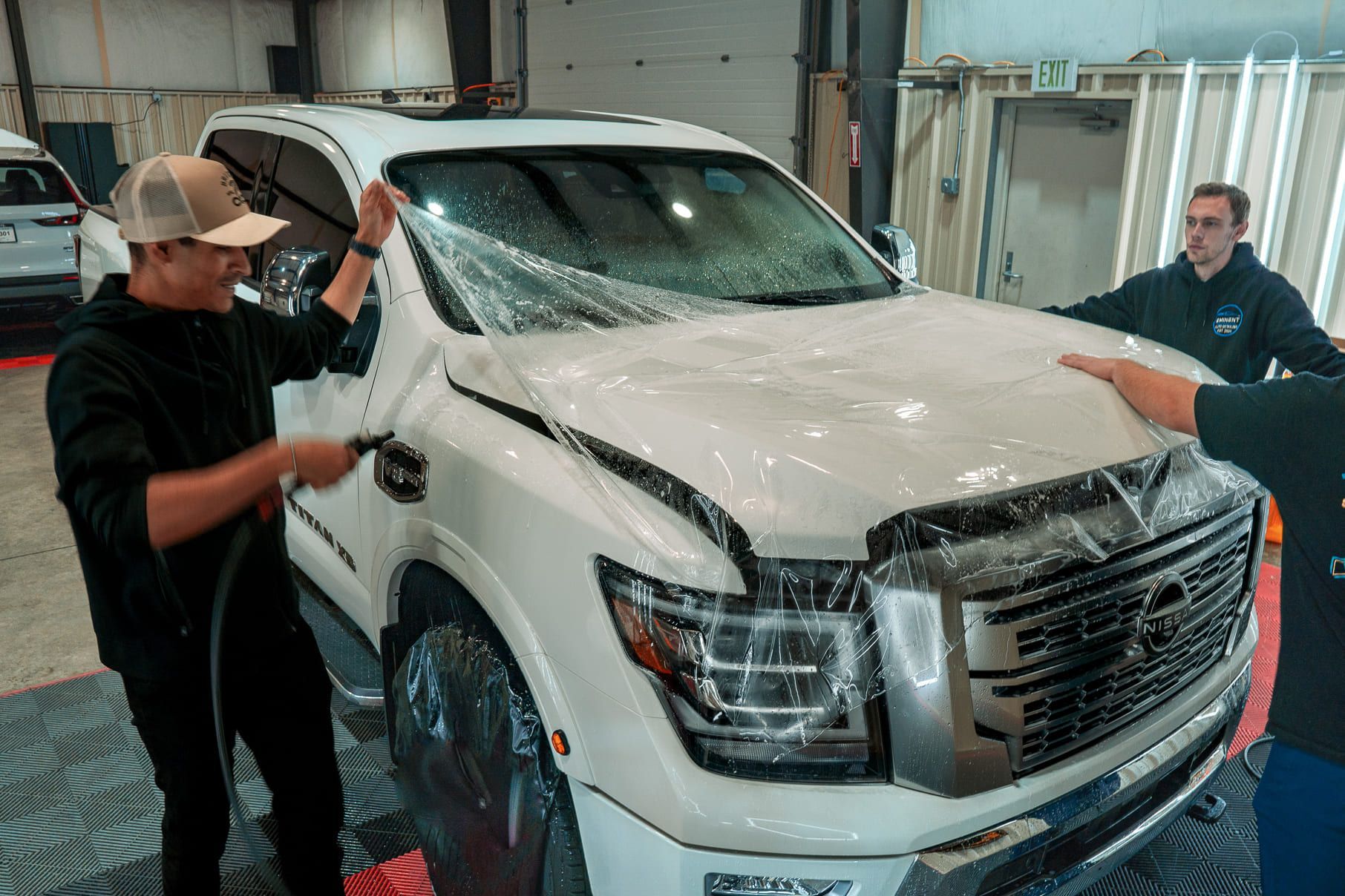 Three people applying protective film to the hood of a white SUV in a workshop. One sprays water.