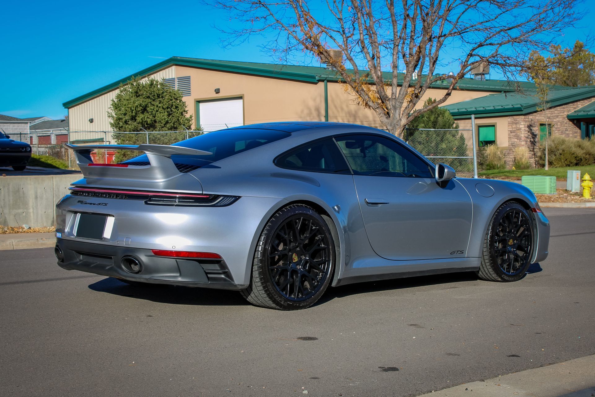 Silver Porsche 911 sports car parked on asphalt, black wheels, rear view, building backdrop.