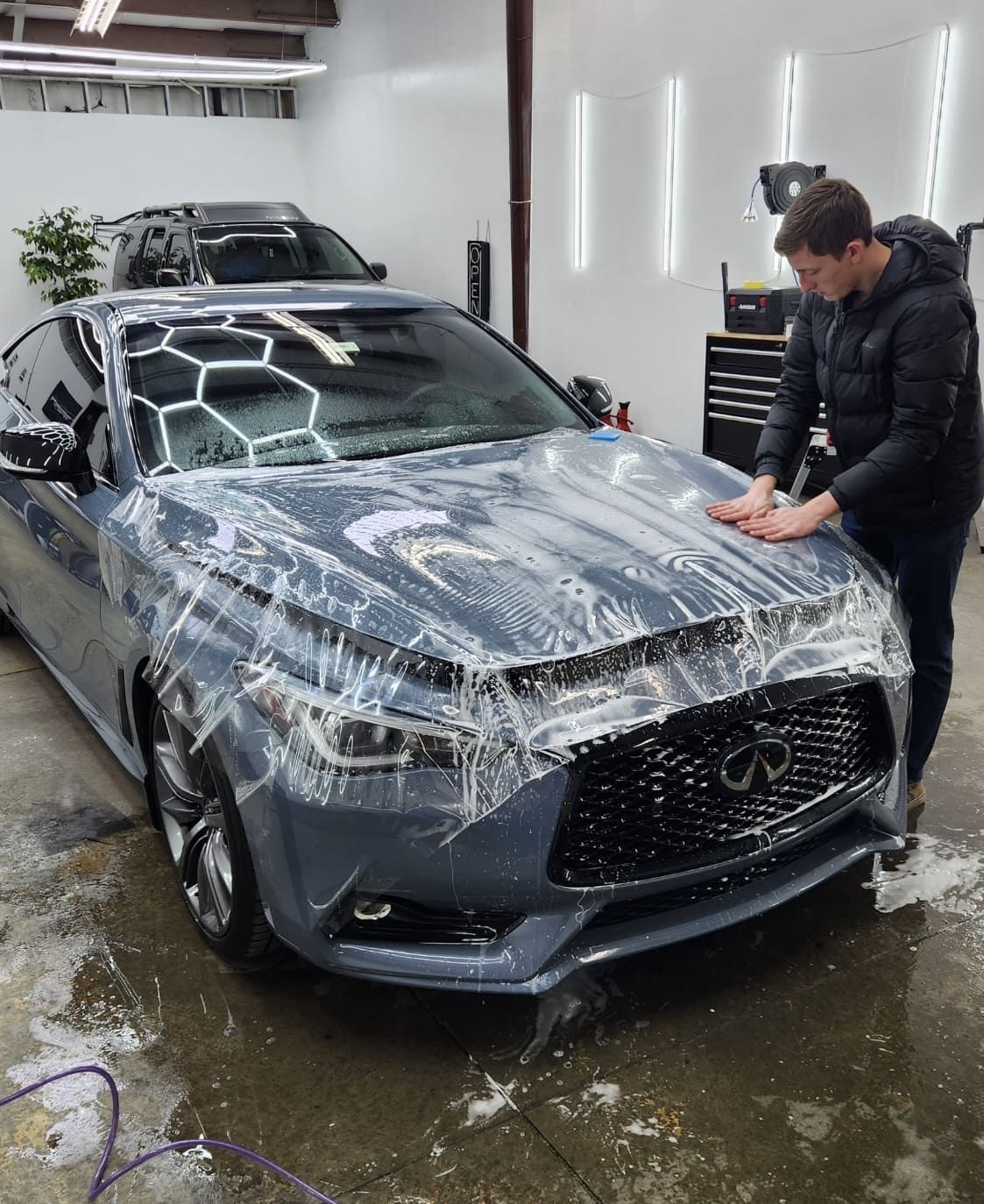 Man applying protective film to a gray Infiniti car in a garage.
