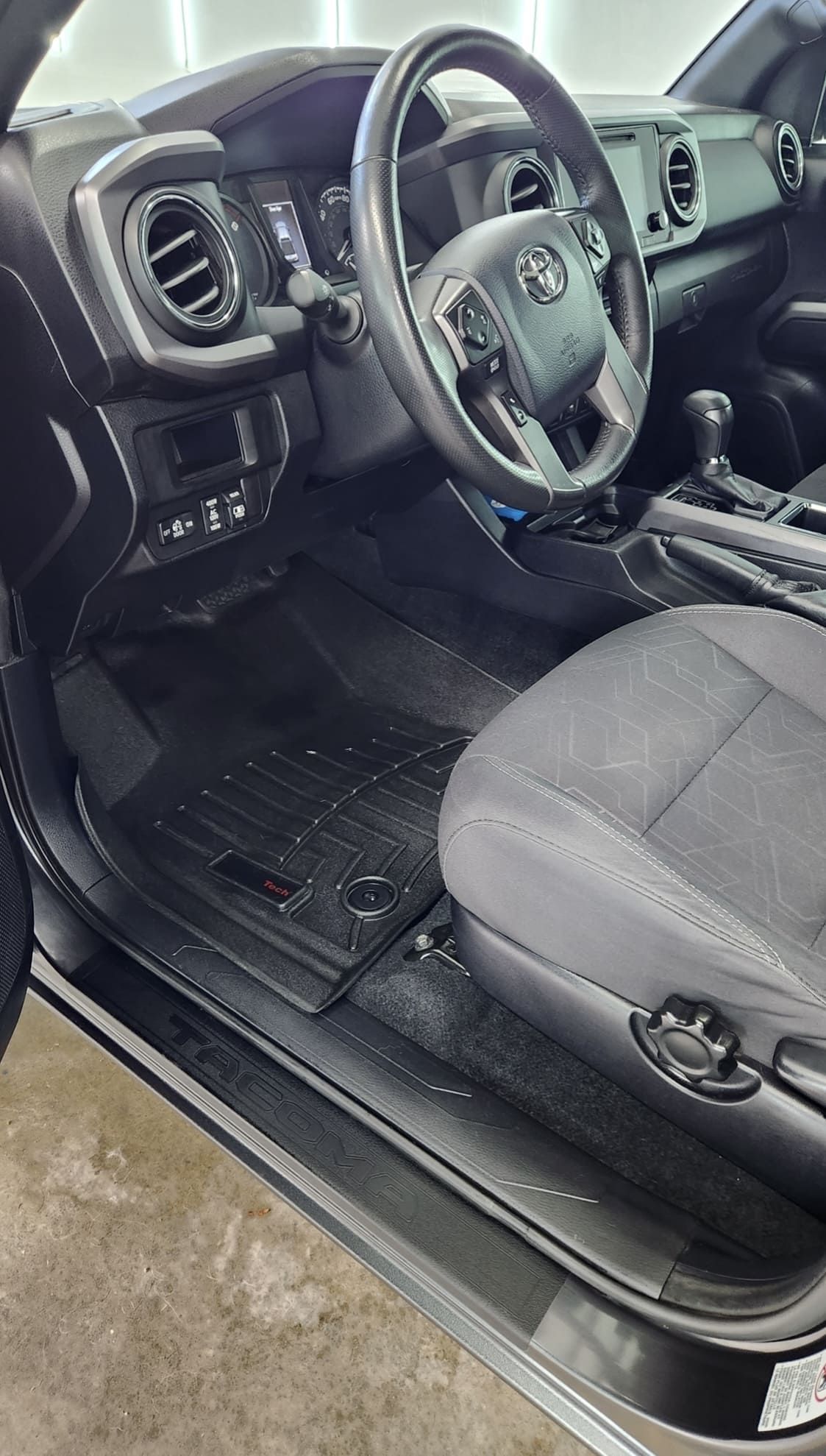 Interior of a black Toyota Tacoma truck with a black steering wheel, dashboard, and floor mats.