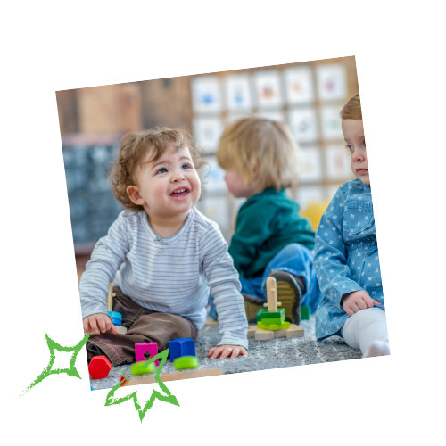 Three toddlers playing at a table indoors: one smiling, another in green, the third in blue.