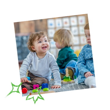 Three toddlers playing at a table indoors: one smiling, another in green, the third in blue.