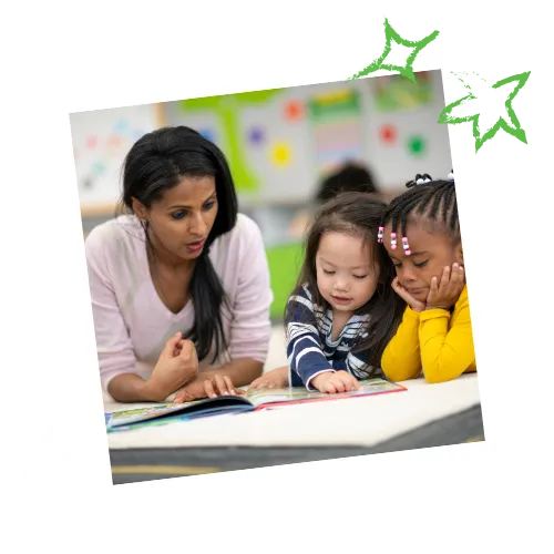 A teacher reads a book with two young students at a table. The children are diverse and focused on the book.