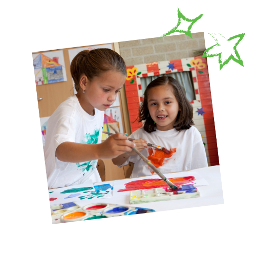 Two young girls painting together at a table, one guiding the other's brush. They are in a classroom.