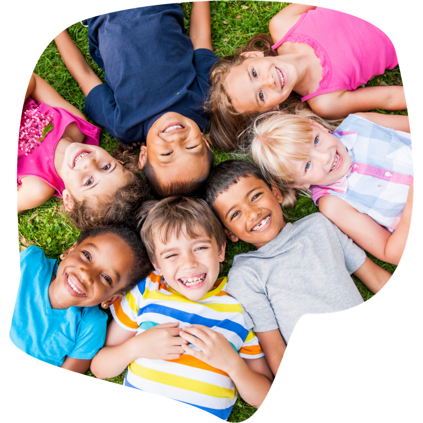 Group of smiling children of diverse ethnicities lying on grass in a circle, looking up.