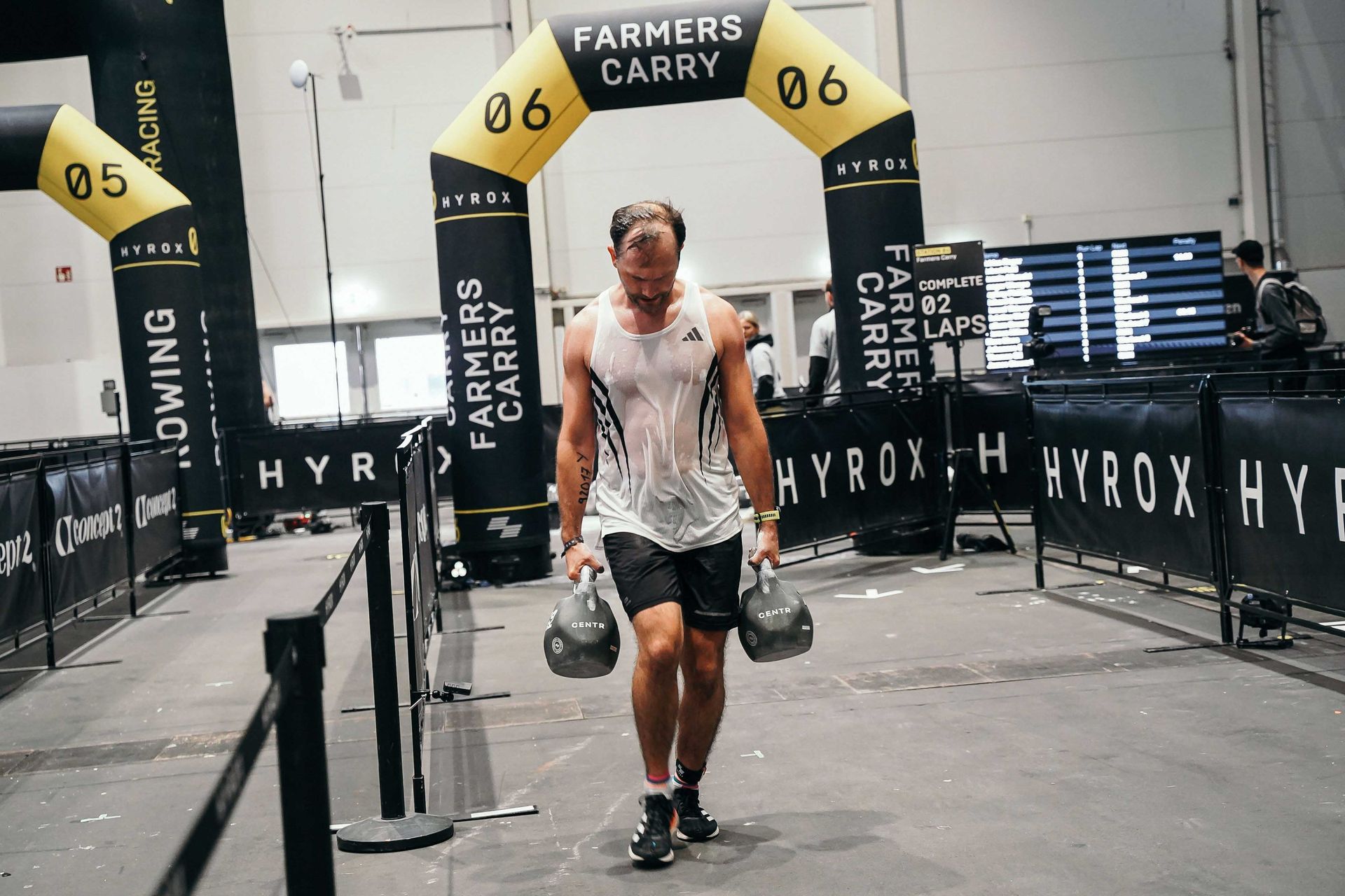 Ein Mann im durchgeschwitzten weißen Tanktop bei der HYROX Übung Farmers Carry