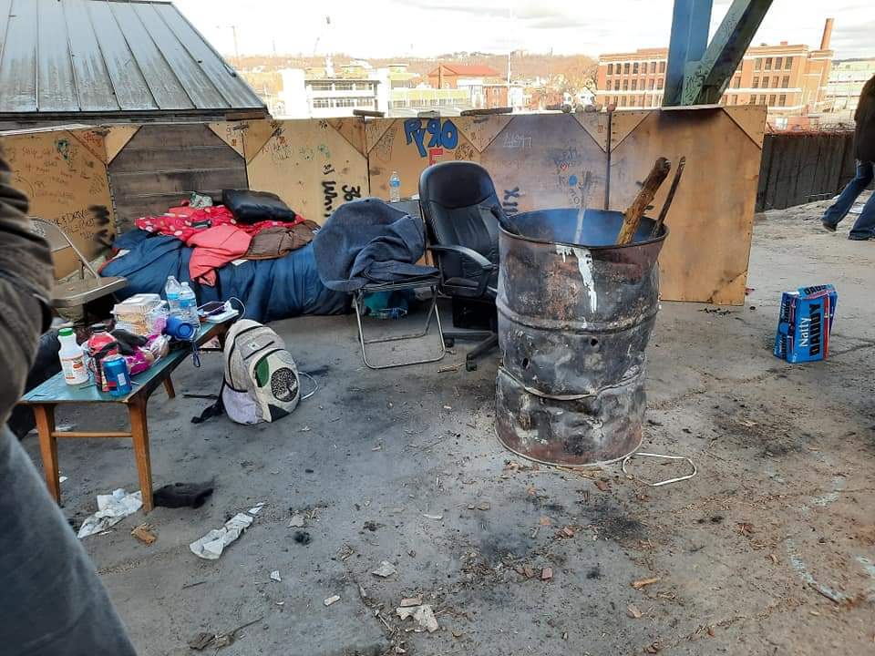A person is standing in a messy room with a barrel and a chair.