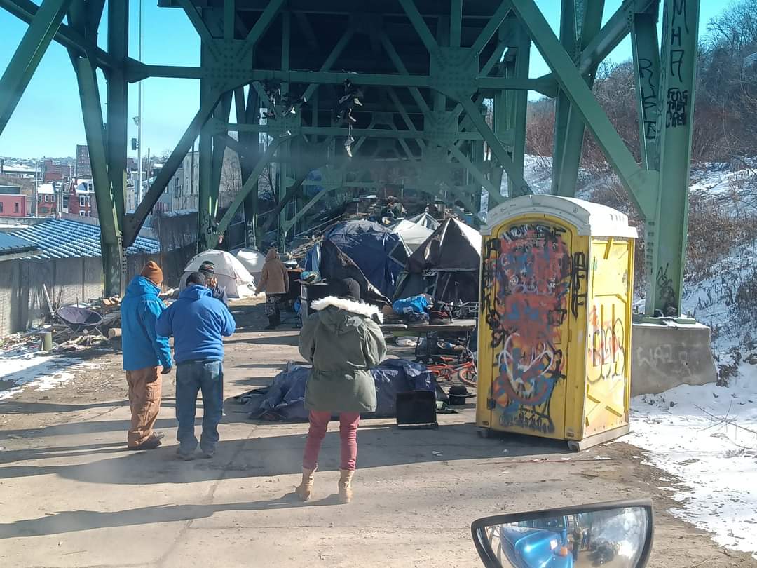 A group of people standing under a bridge next to a portable toilet with graffiti on it