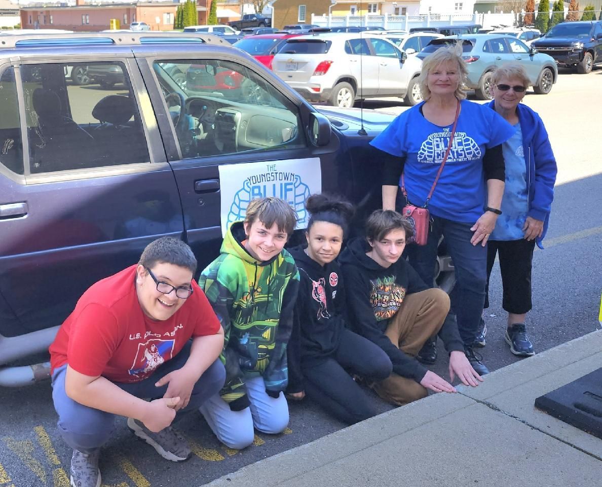 A group of people are posing for a picture in front of a car with a sign on the door that says